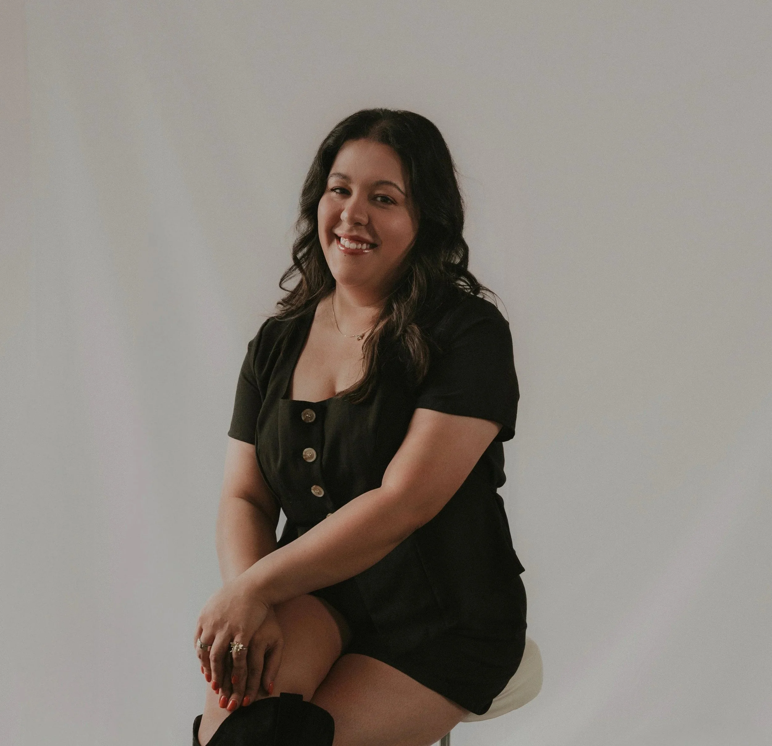 A woman with dark hair, wearing a black dress with buttons, sitting on a stool against a plain white wall, smiling and looking at the camera.