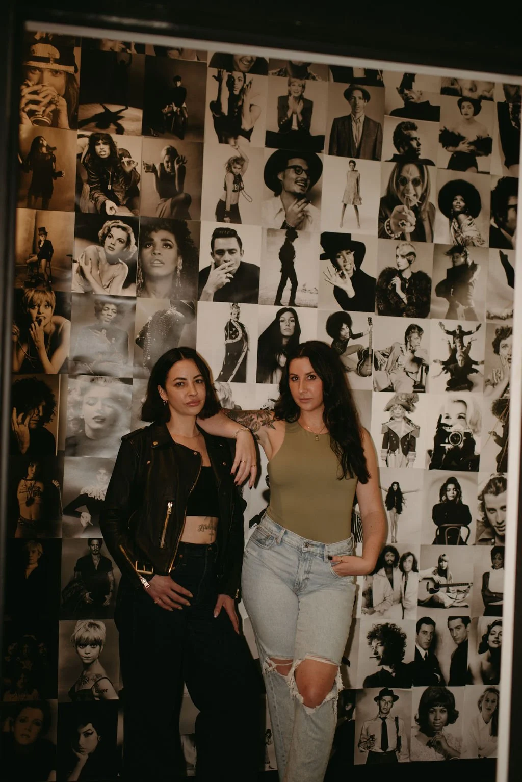 Two women posing in front of a black and white collage of celebrity photos on a wall, one wearing a leather jacket and the other in a tank top and jeans.