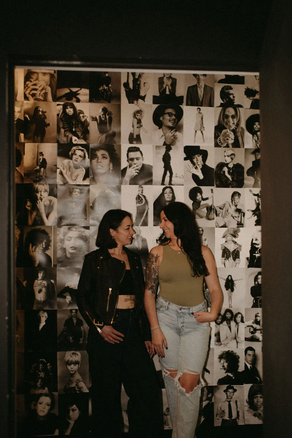 Two women standing in front of a collage of black-and-white celebrity photos, smiling at each other. One woman is wearing a black jacket and pants, the other a green tank top and ripped jeans.