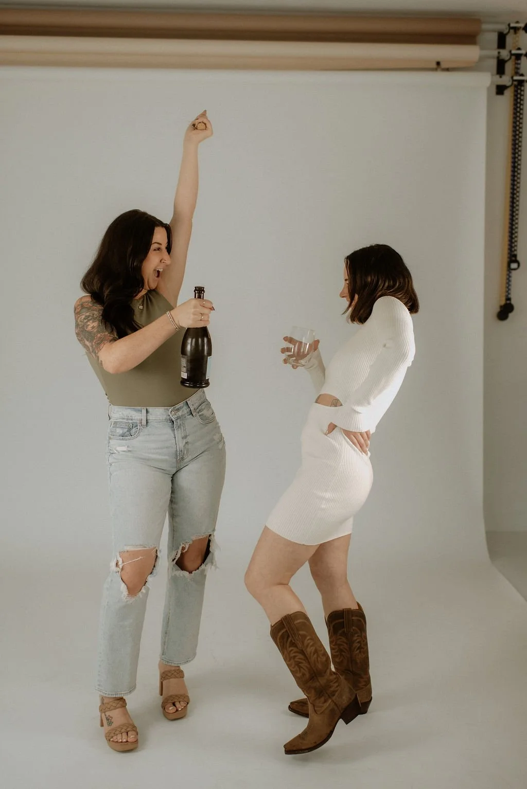 Two women having fun, one holding a bottle, the other with a glass, in a studio setting.