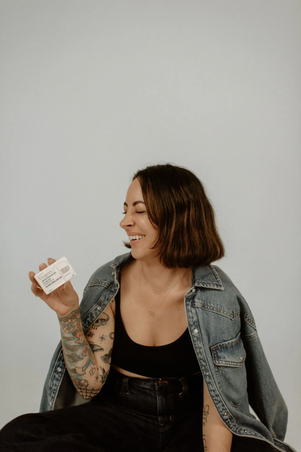 A person with tattooed arms holding a product box, wearing a denim jacket and black top, smiling against a plain background.