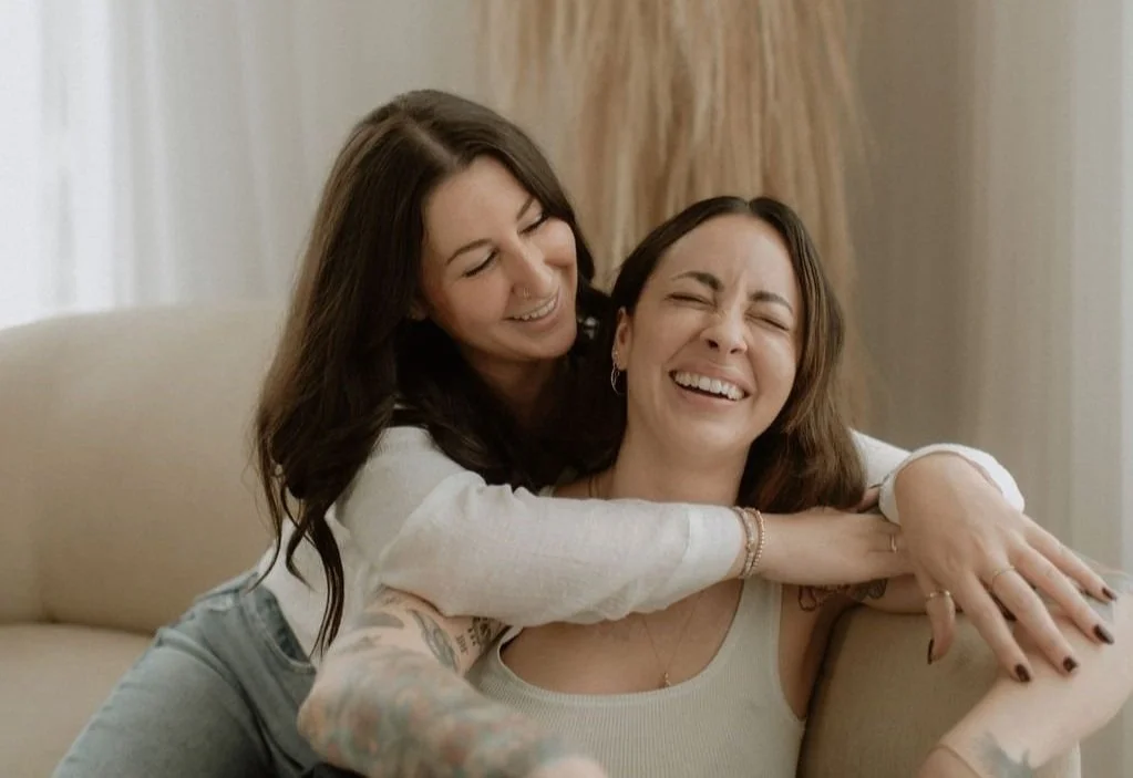 Two women laughing and hugging on a sofa in a bright room, with one woman having tattoos on her arm.