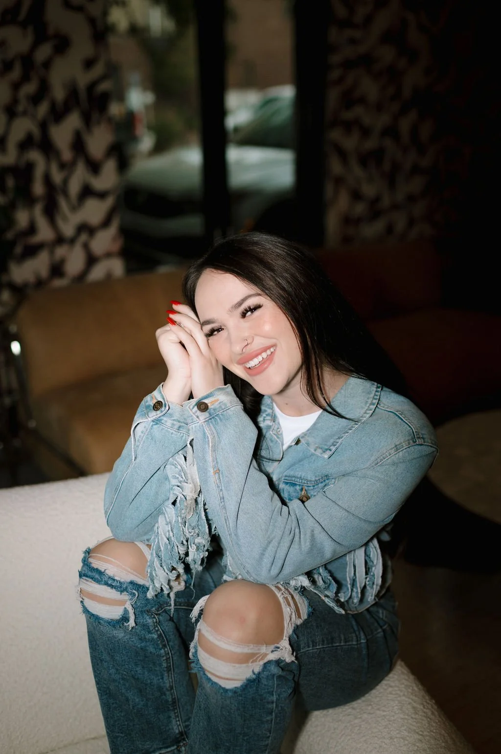 Woman smiling in a denim jacket and ripped jeans, sitting indoors.
