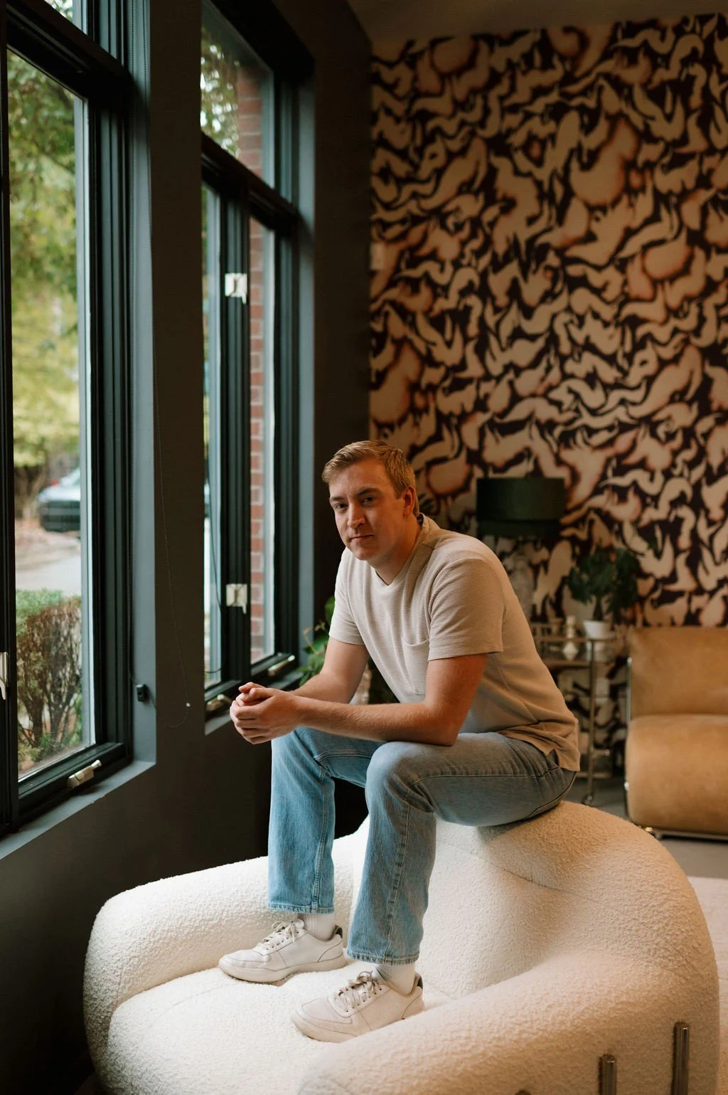 Man sitting on a modern white armchair near large windows with a patterned wall in the background.