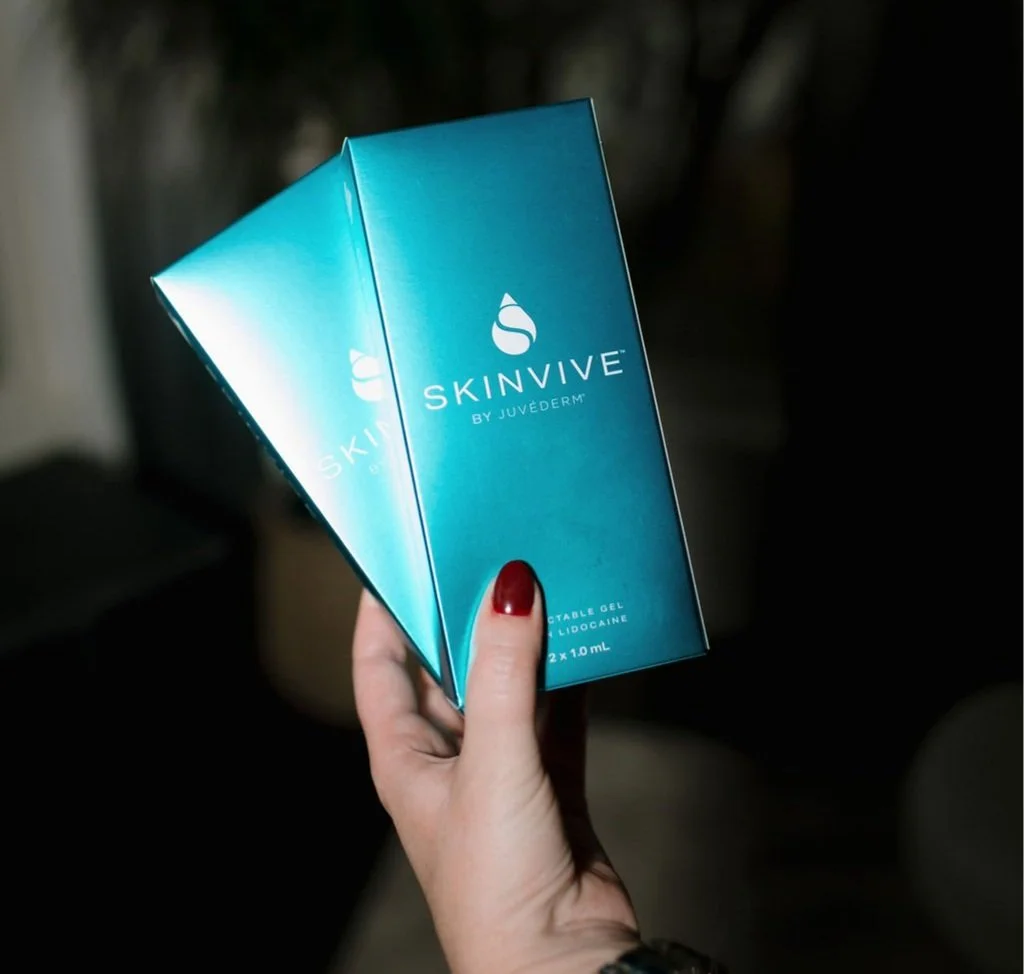 A hand with red nail polish holding two blue boxes of SkinVive by Juvederm, containing injectable gel with lidocaine, in a dimly lit environment.