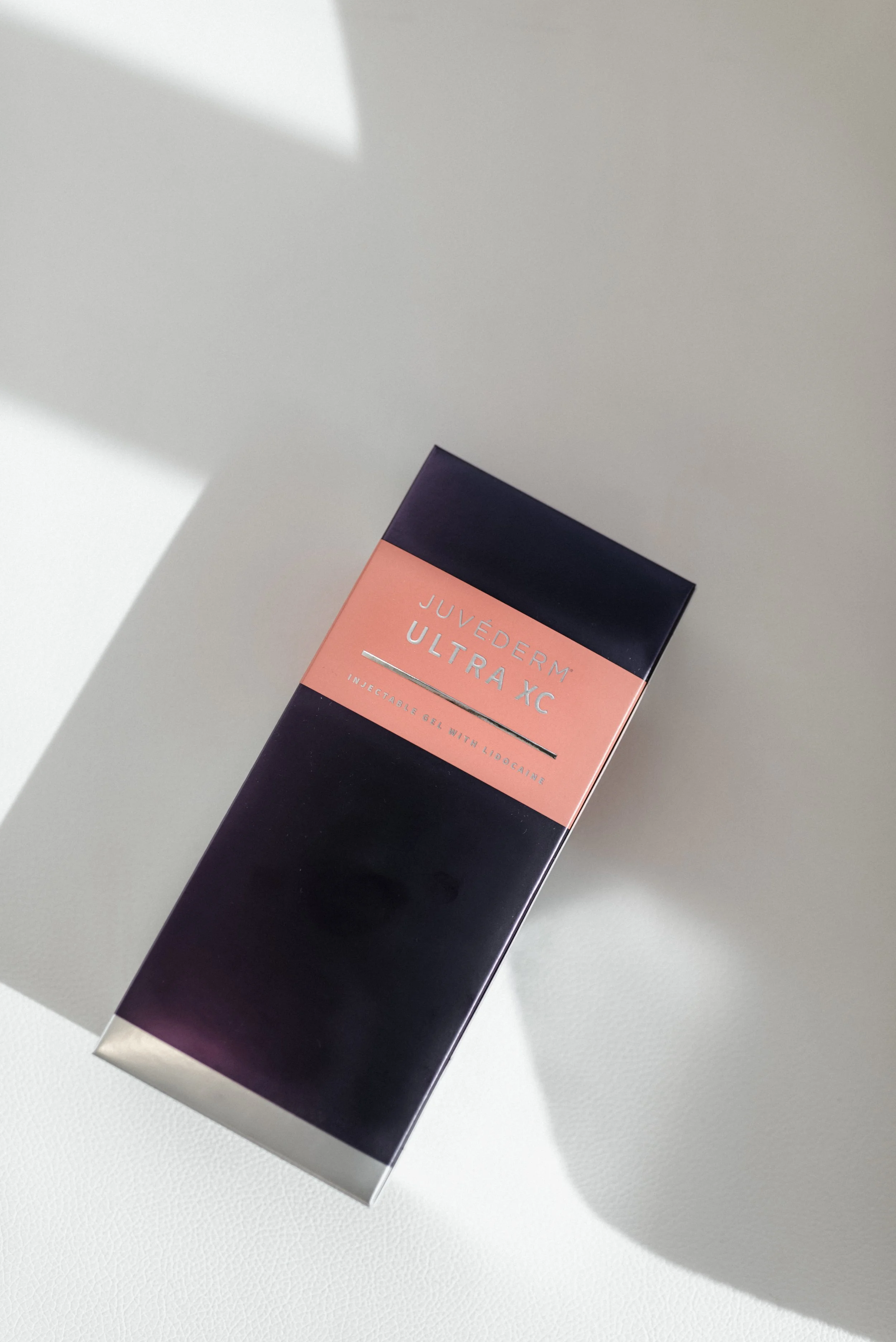 Close-up of a Juvederm Ultra XC injectable gel box with pink and dark blue packaging on a white surface, casting a shadow.