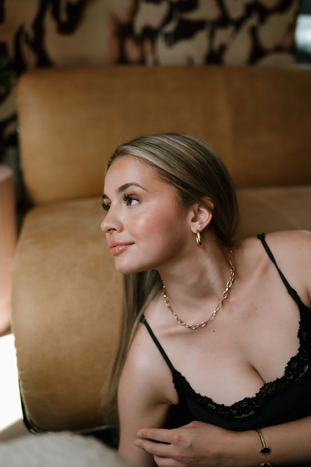 A woman with long brown hair lying on a couch, wearing a black lace camisole, gold hoop earrings, a gold chain necklace, and a gold bracelet, looking to the left with a subtle smile.