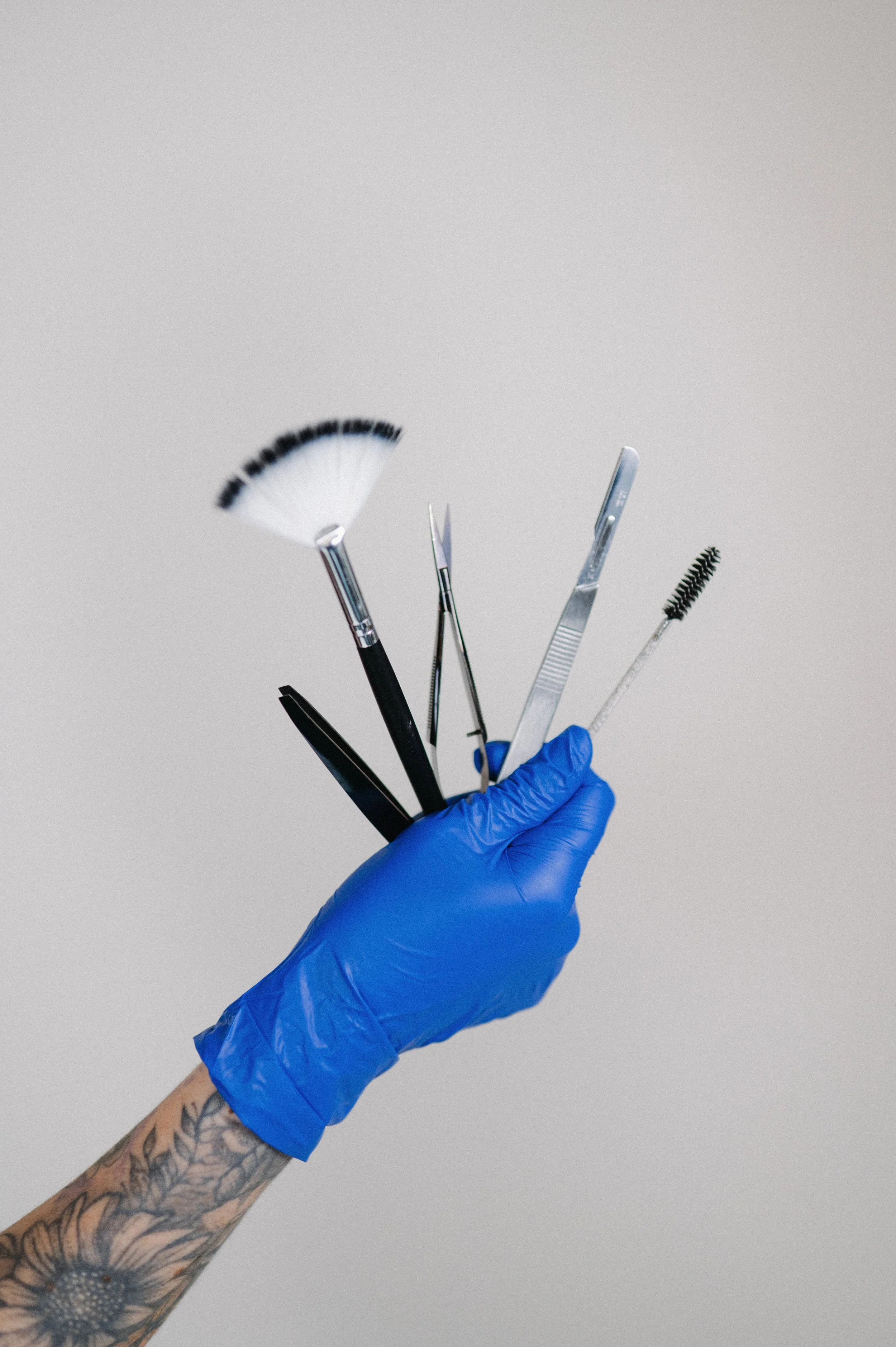 A hand with tattoos wearing a blue glove holding a collection of tattooing and microblading tools, including brushes, scissors, tweezers, and other metal instruments against a plain grey background.