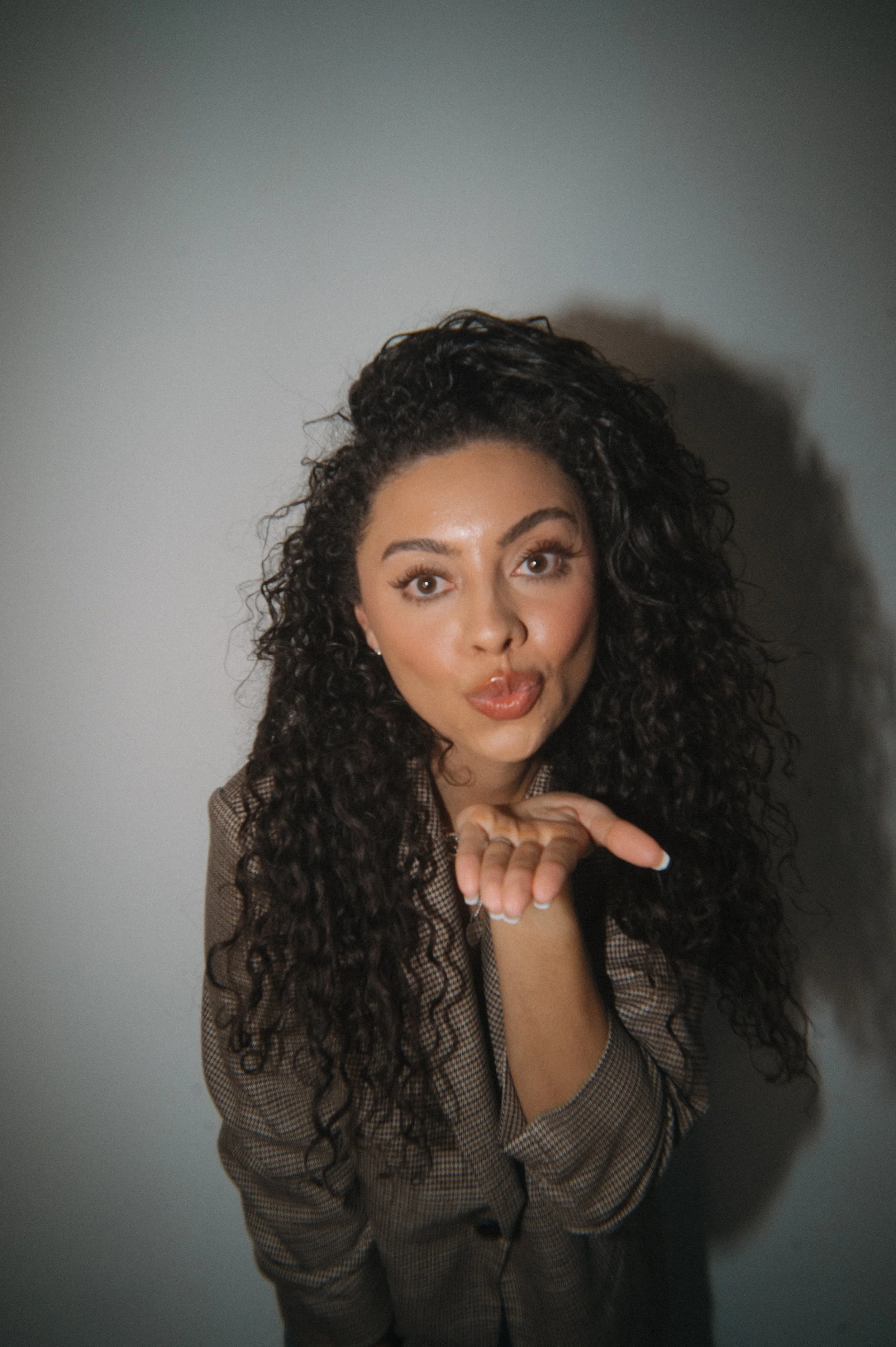 A woman with long curly hair, wearing a checkered blazer, is making a playful gesture with her hand near her face. She is looking directly at the camera against a plain grey background.