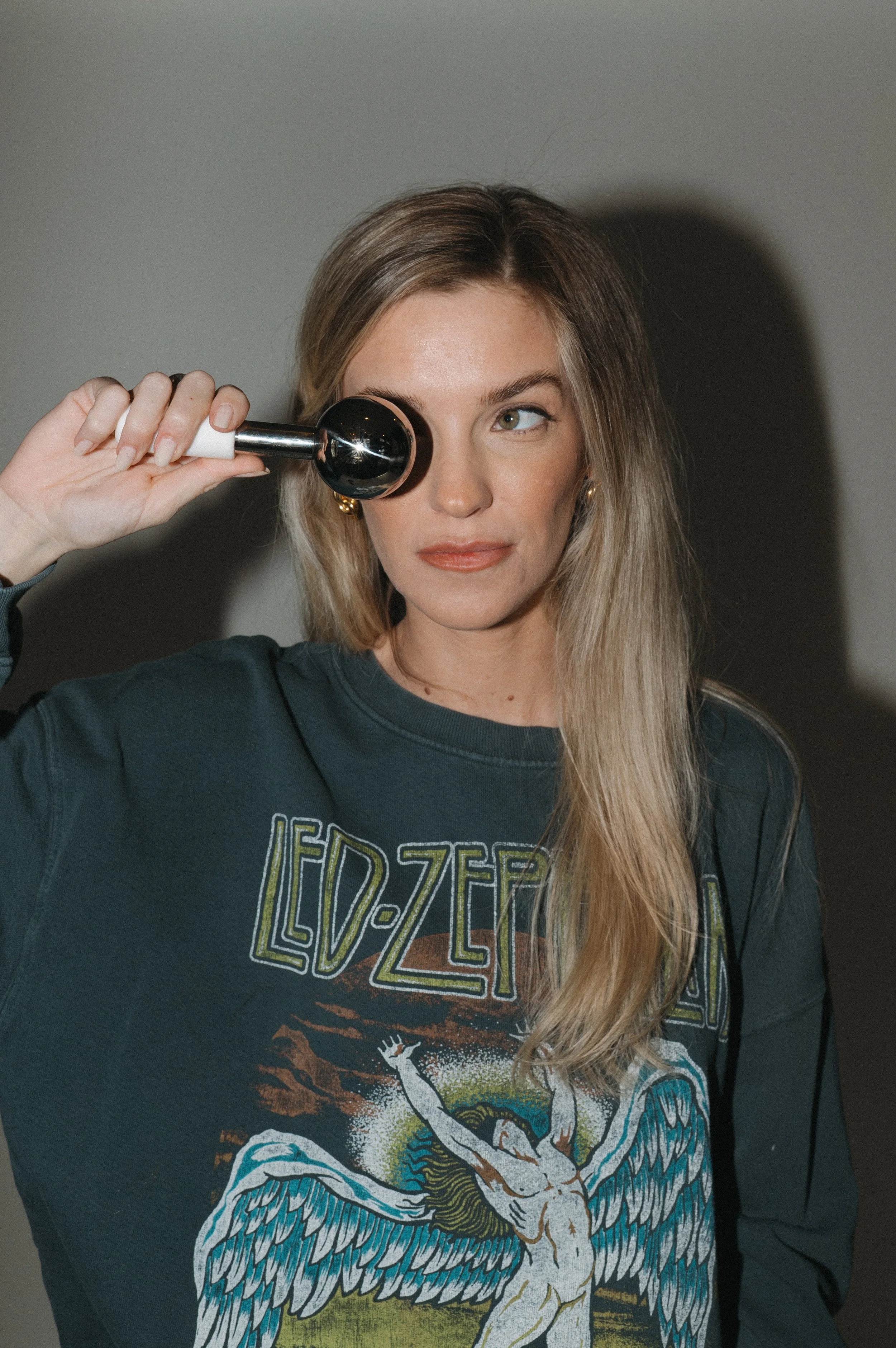 A woman with light brown hair and earrings holds a metal and white beauty tool to her face, looking to the side with a slight smile, wearing a Led Zeppelin T-shirt.