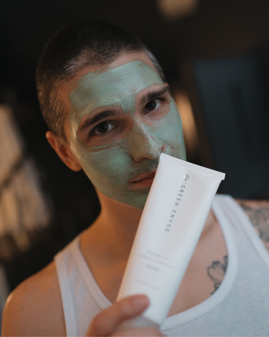 A young man with short dark hair and a sleeveless white shirt applies a green facial mask, holding a tube of skincare product labeled 'Green Envie' in front of him.
