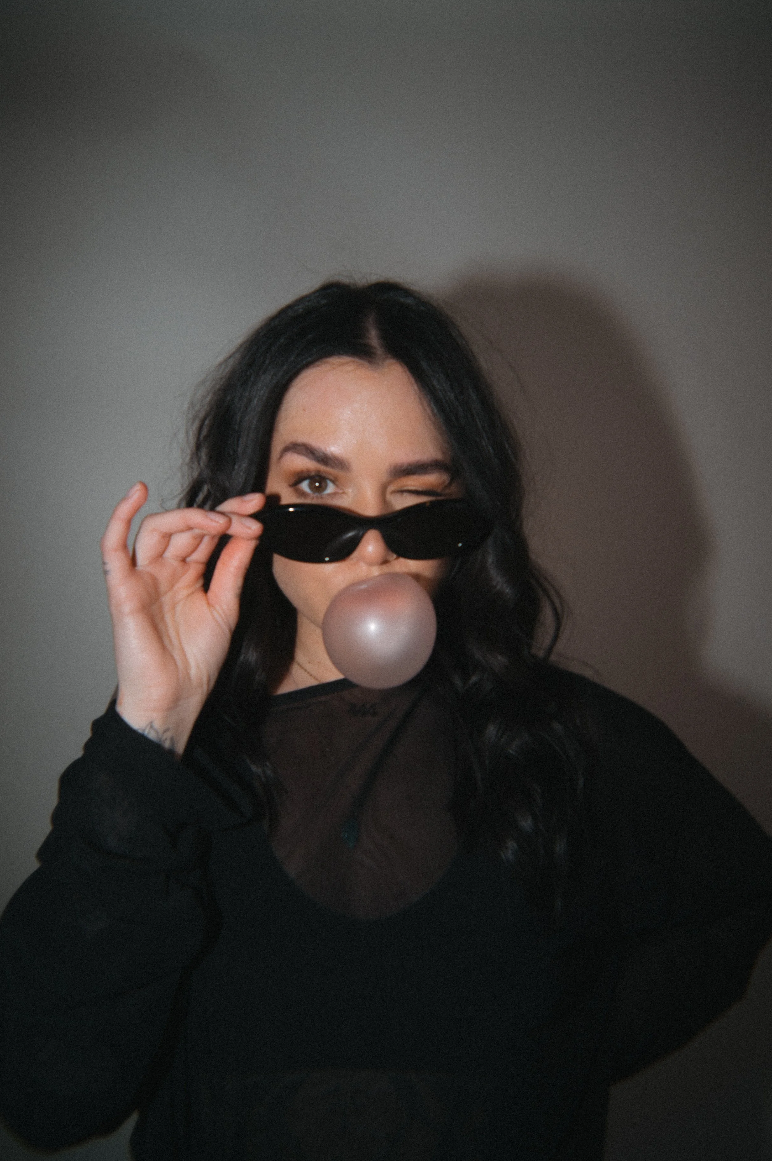A woman with long black hair and tattoos on her hand, wearing black sunglasses and a black top, is holding the sunglasses with her right hand and blowing a pink bubble gum.