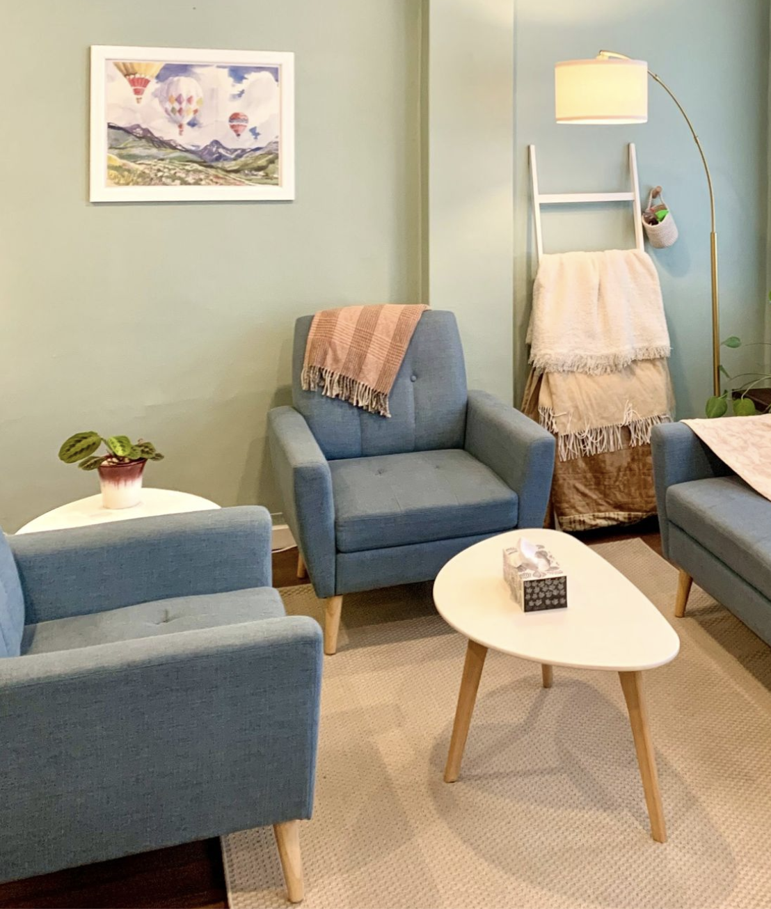 Living room with blue armchairs, a white oval coffee table with a tissue box, a framed landscape painting, a potted plant, and a floor lamp with a white shade. A blanket is draped over the back of one armchair, and a blanket hangs on a ladder-style blanket rack.