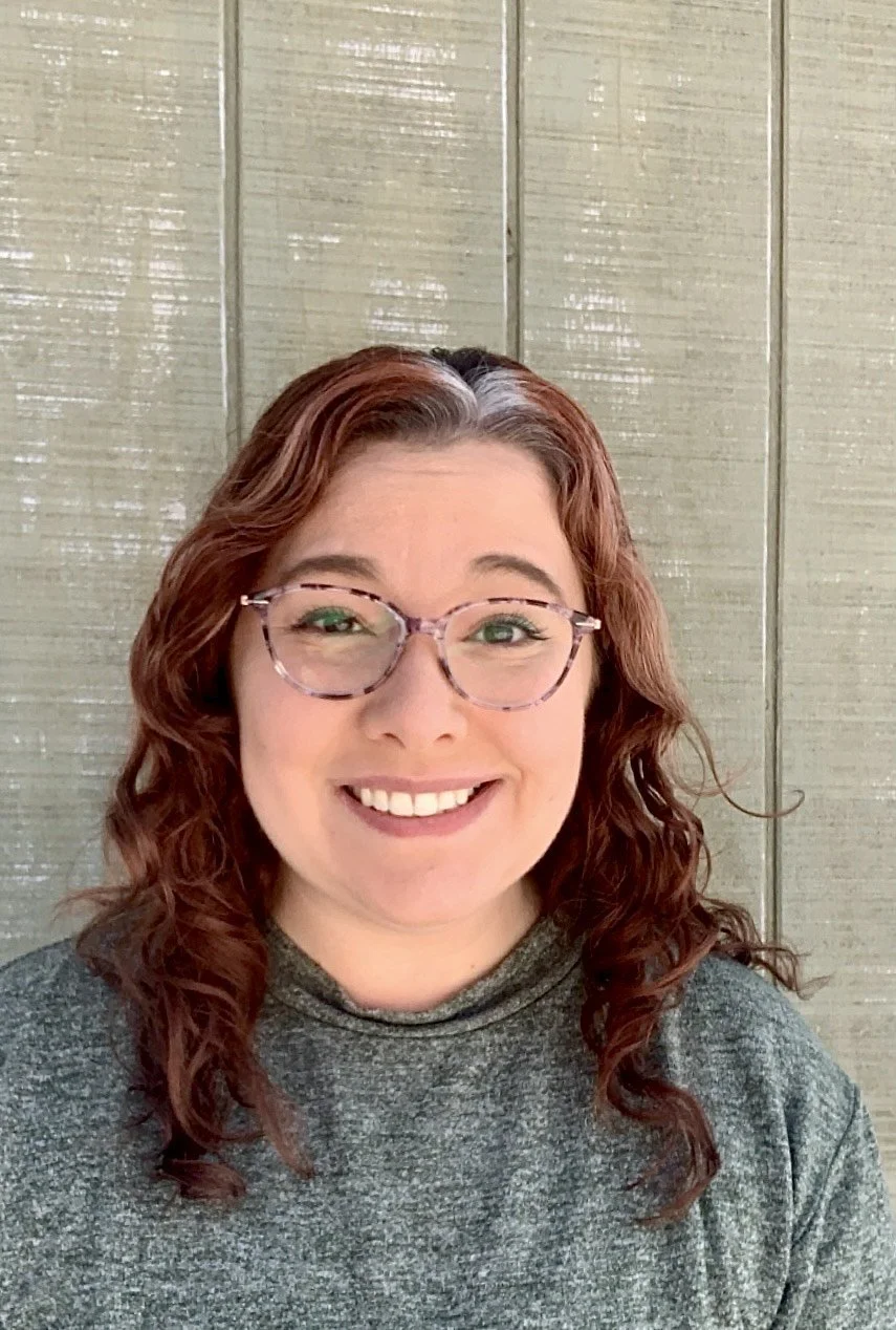 A woman with curly auburn hair, wearing glasses with a tortoiseshell pattern and a gray turtleneck sweater, smiling in front of a wooden background.