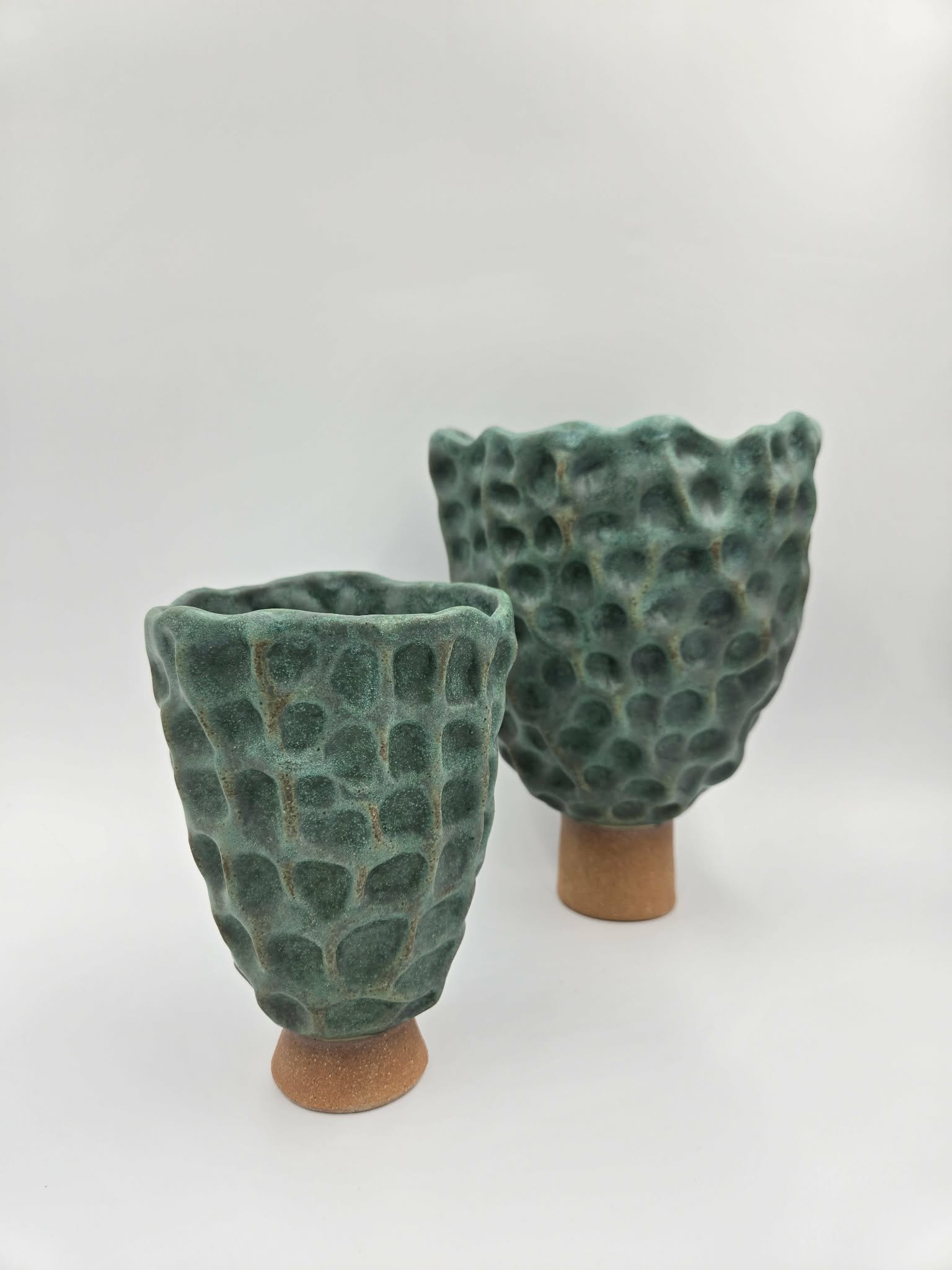 Deux vases en céramique vert à motif de galets, posés sur des supports en terre cuite, sur un fond blanc.