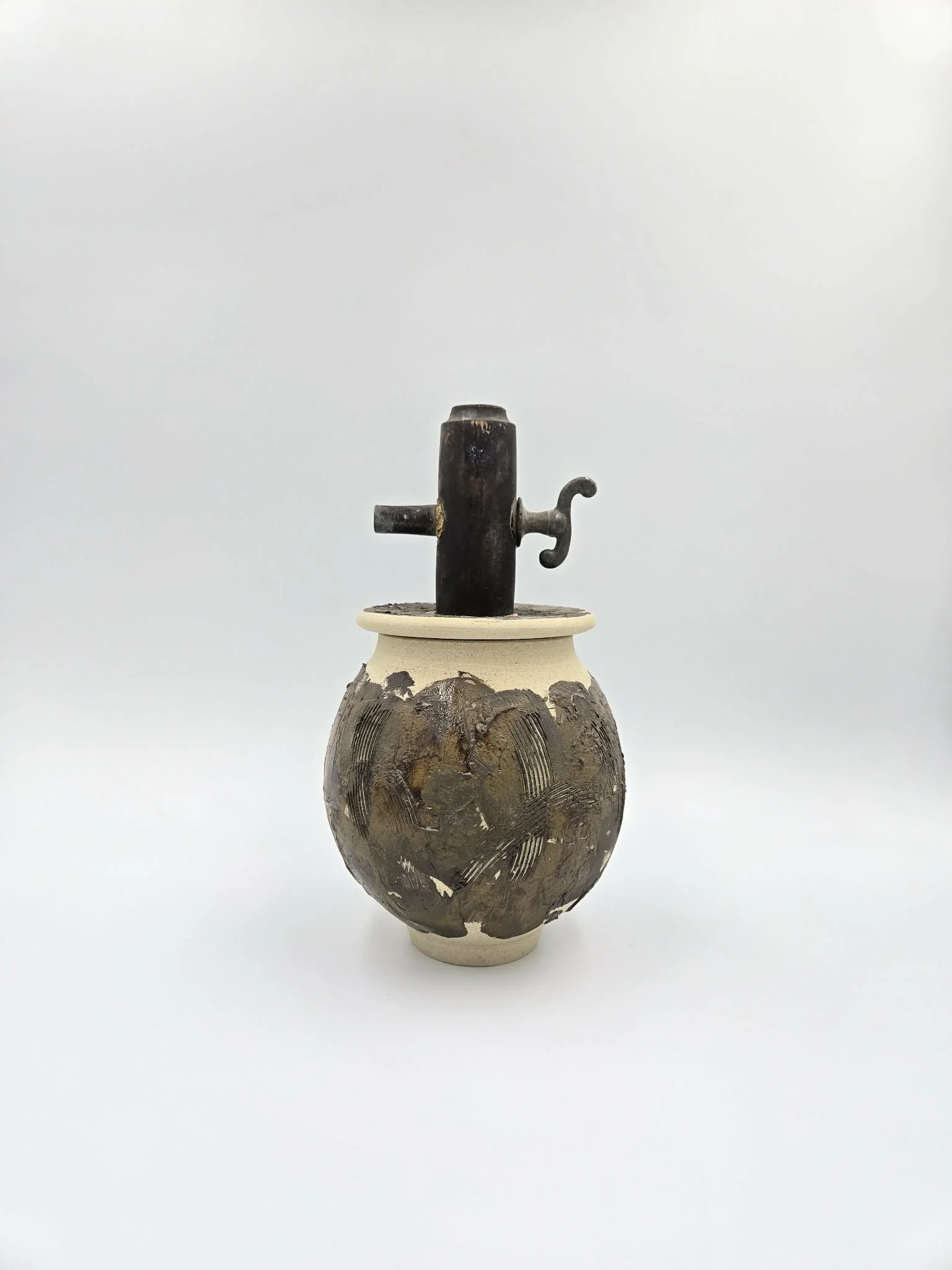 Une jarre en céramique avec une surface décorative, posée sur une surface blanche. Surmontée d'un objet métallique noir en forme de valve ou robinet, doté d'une poignée en forme de « S ».
