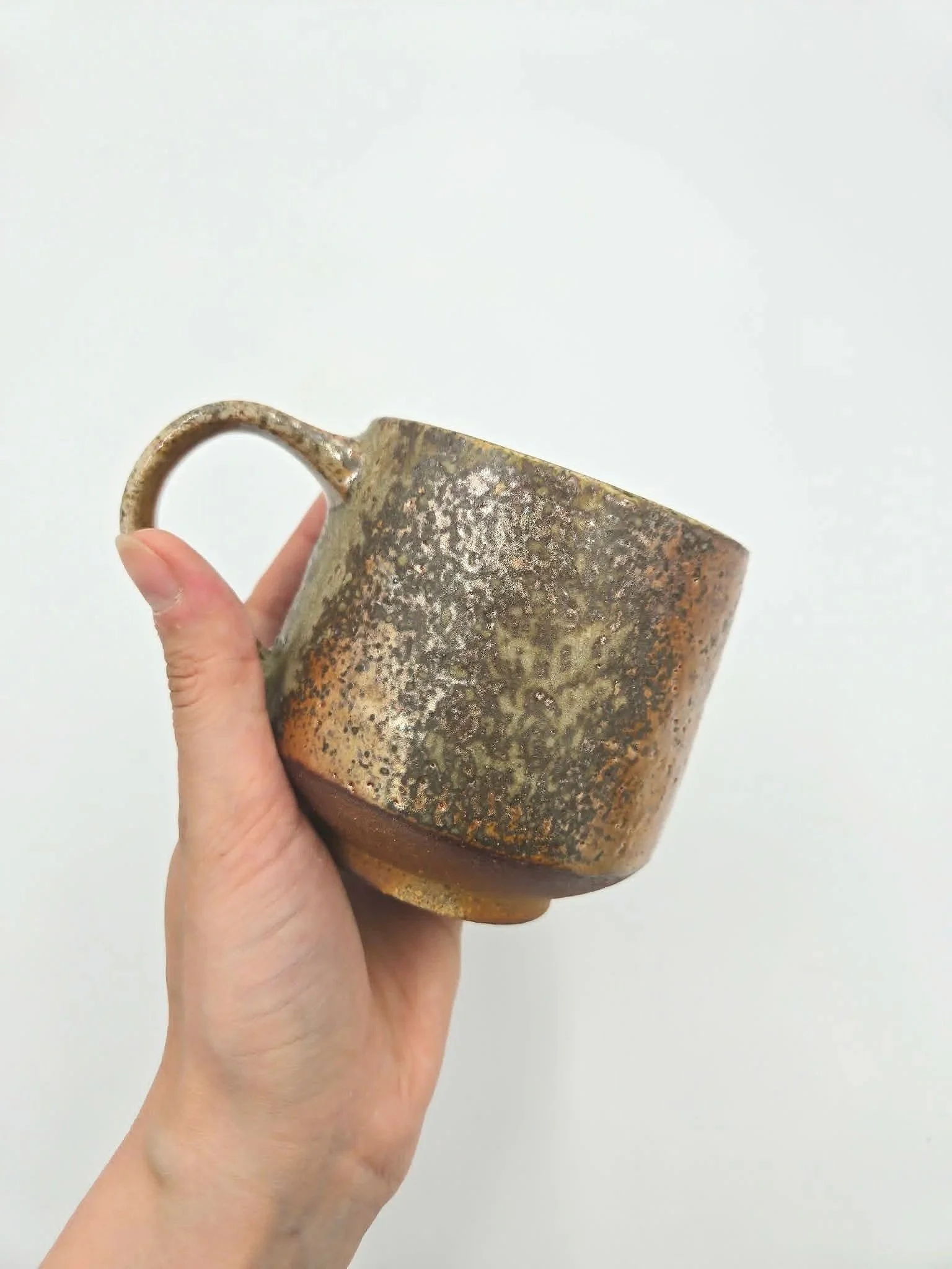 Tasse en céramique cuite au four a bois avec une surface texture rustique, tenue par une main contre un fond blanc.