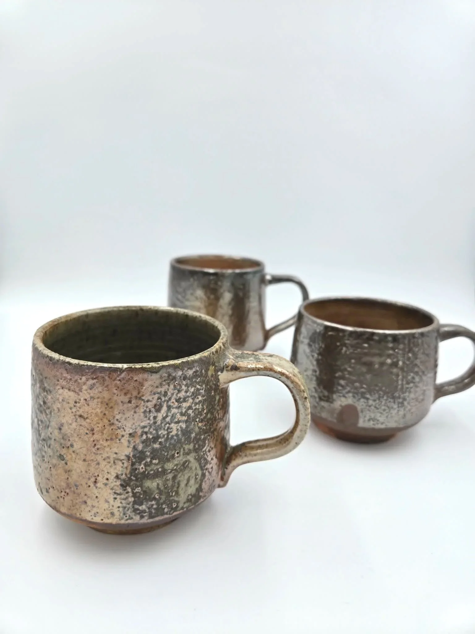 Trois mugs en céramique artisanale avec une finition rustique et une glaçure brillante, disposés sur une surface blanche.