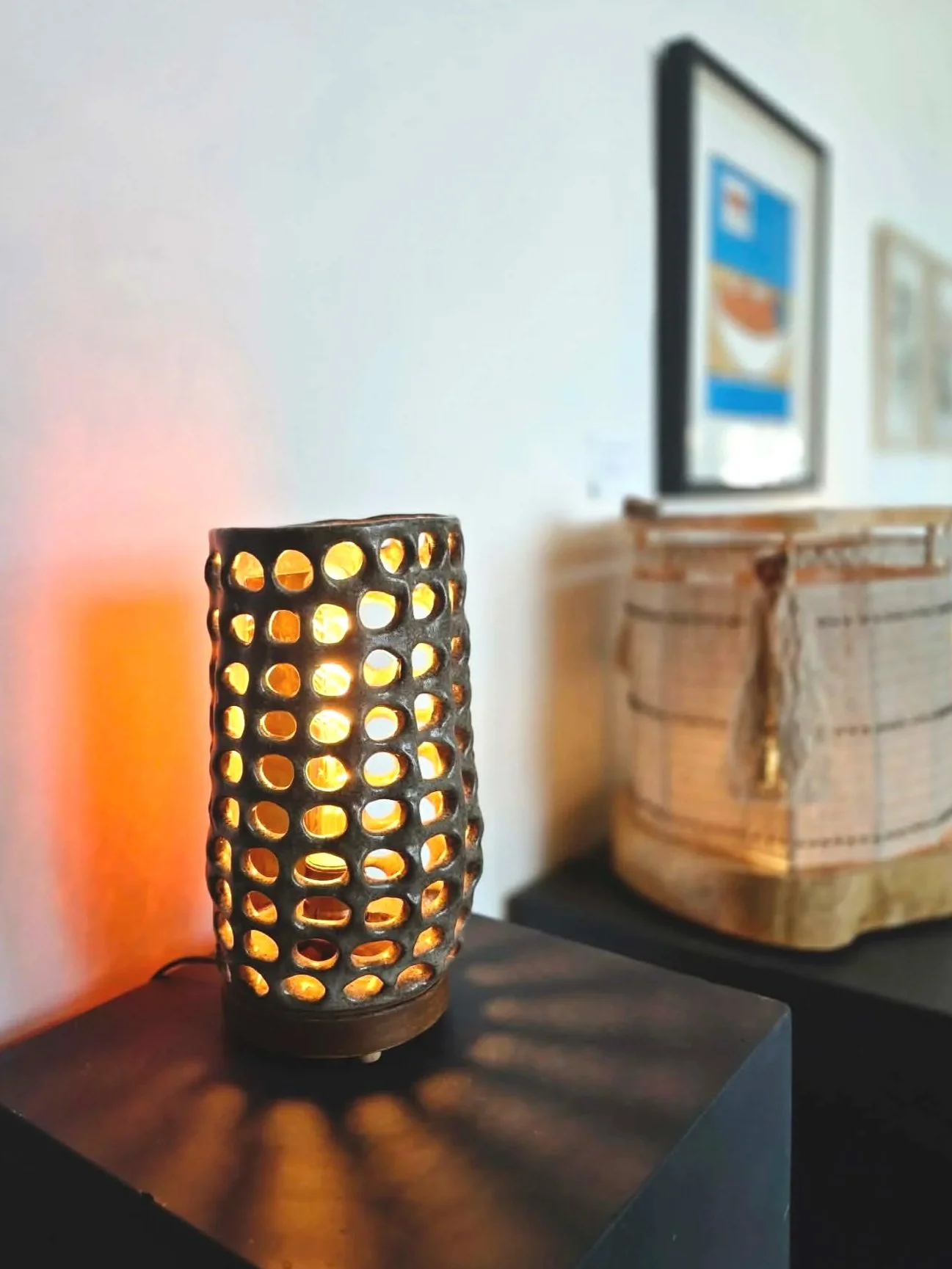 Lampe de table cylindrique en métal avec un motif de cercles, éclairée, posée sur une surface noire, avec une source de lumière chaude à l'intérieur. En arrière-plan, un meuble en bois avec un panier, et à droite, un mur blanc avec un écran ou une im