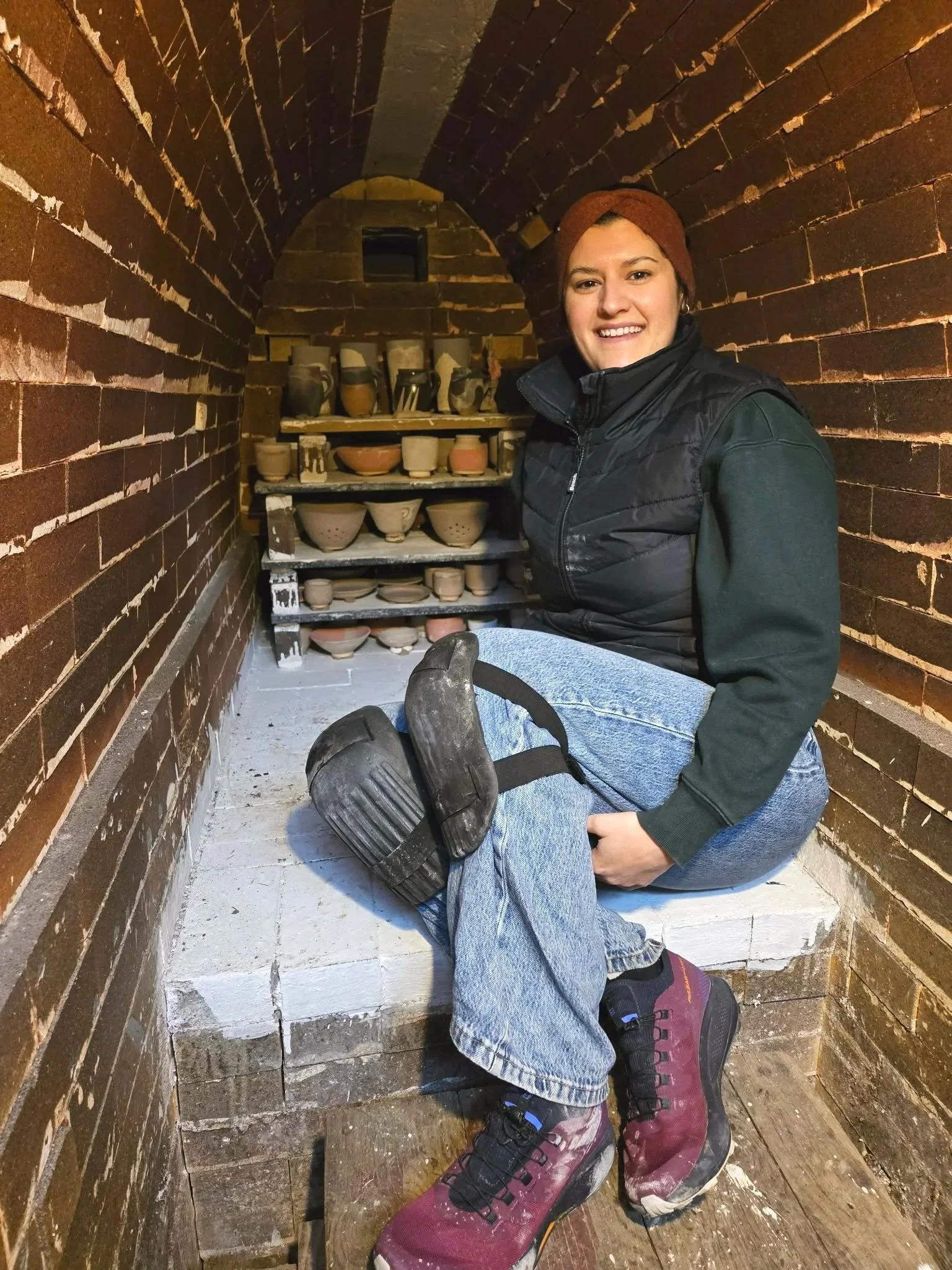 Une femme souriante portant un bonnet marron, une veste noire et des bottes roses, assise dans un espace en pierre ou en brique avec des étagères de poteries derrière elle.