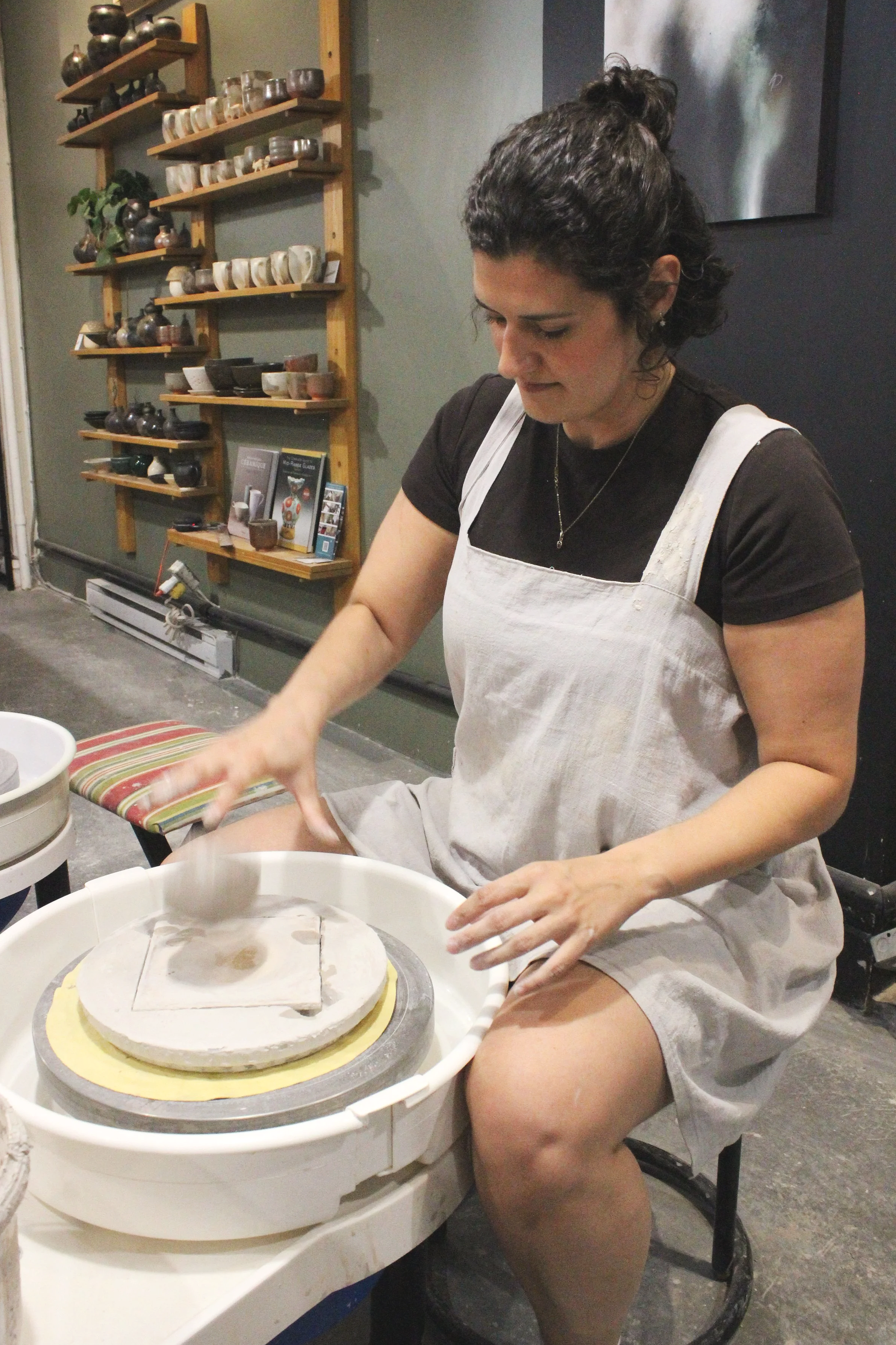 Une femme assis au tour de potier qui lance une boule sur le tour, autour d'elle il y a des tablettes remplis de pièces de poterie
