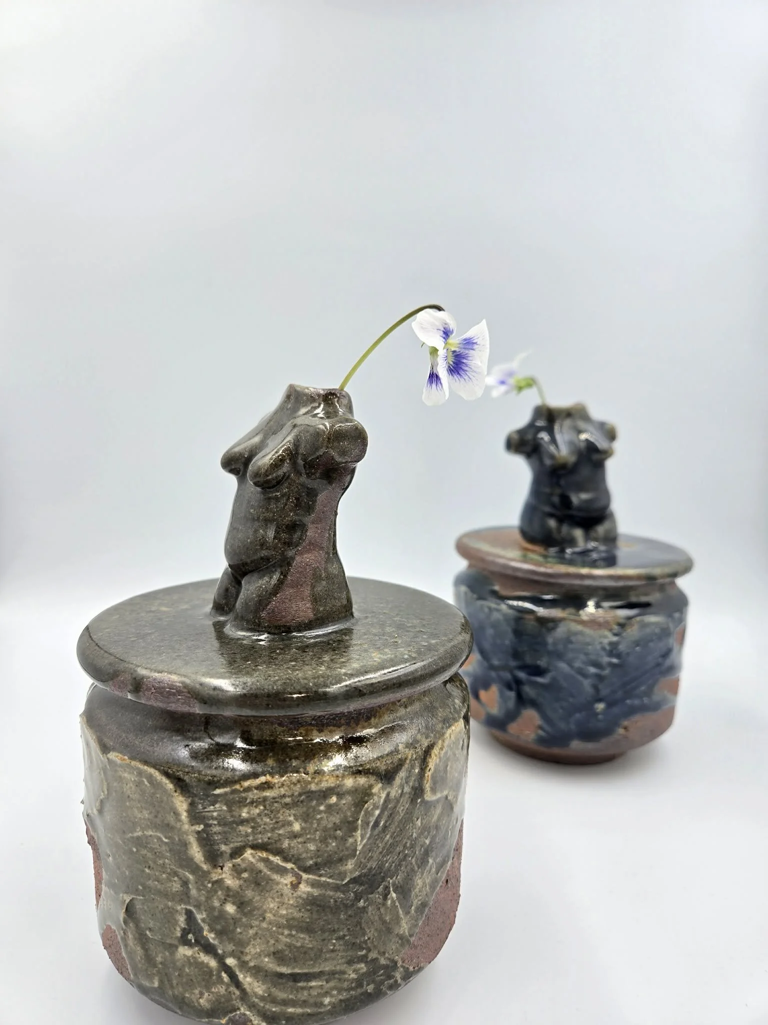 Deux petits vases en poterie avec des motifs en relief, contenant une fleur blanche et violette. Disposés sur une surface blanche.