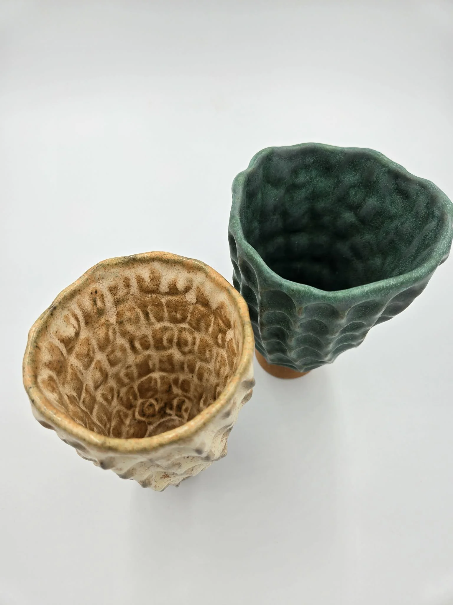 Deux vases en céramique, un beige avec un motif géométrique et un vert foncé avec une texture côtelée, placés sur fond blanc.