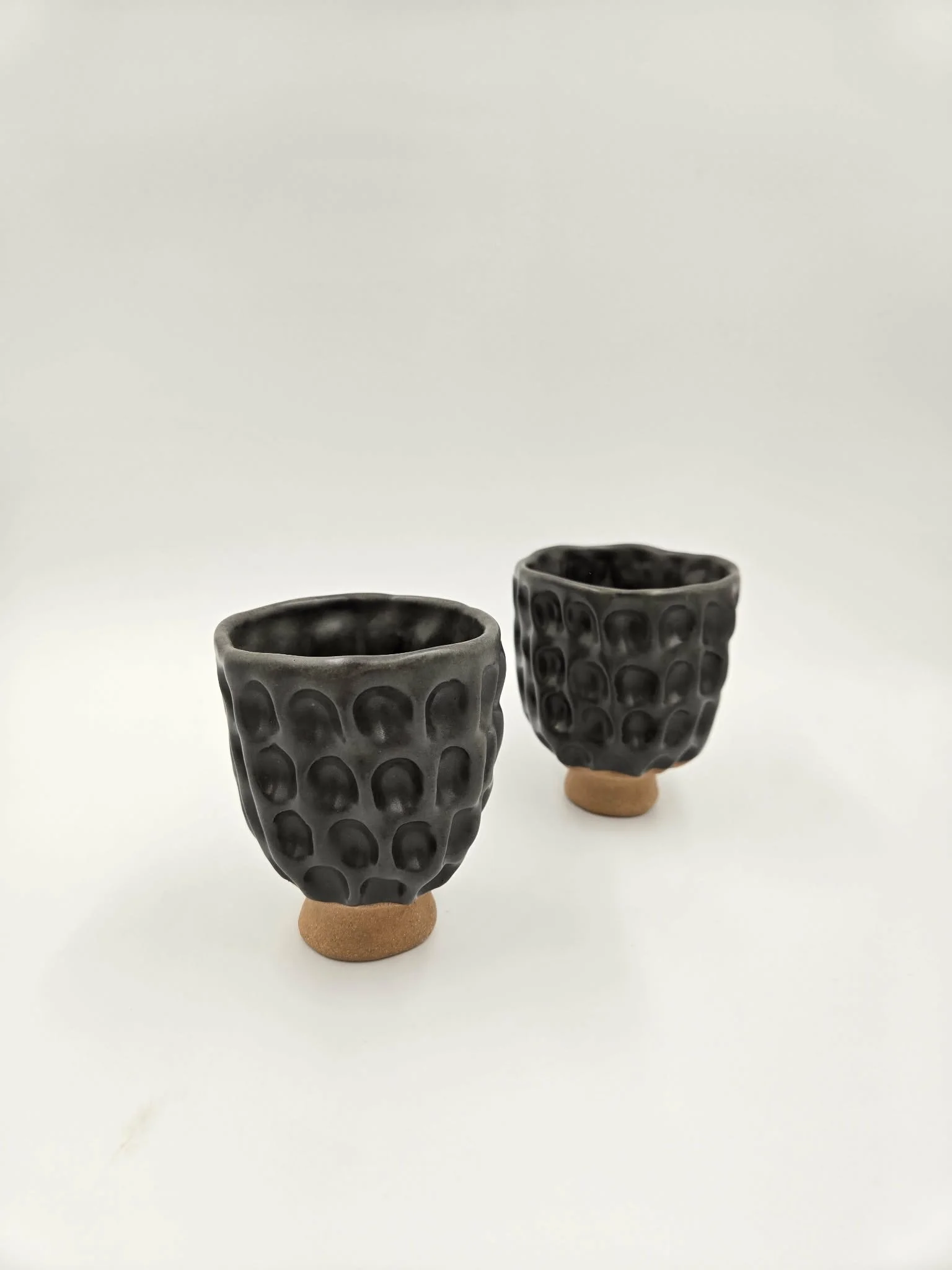 Deux tasses en céramique noire avec une surface texturée de cercles, posées sur de petites bases en terre cuite, contre un fond blanc.