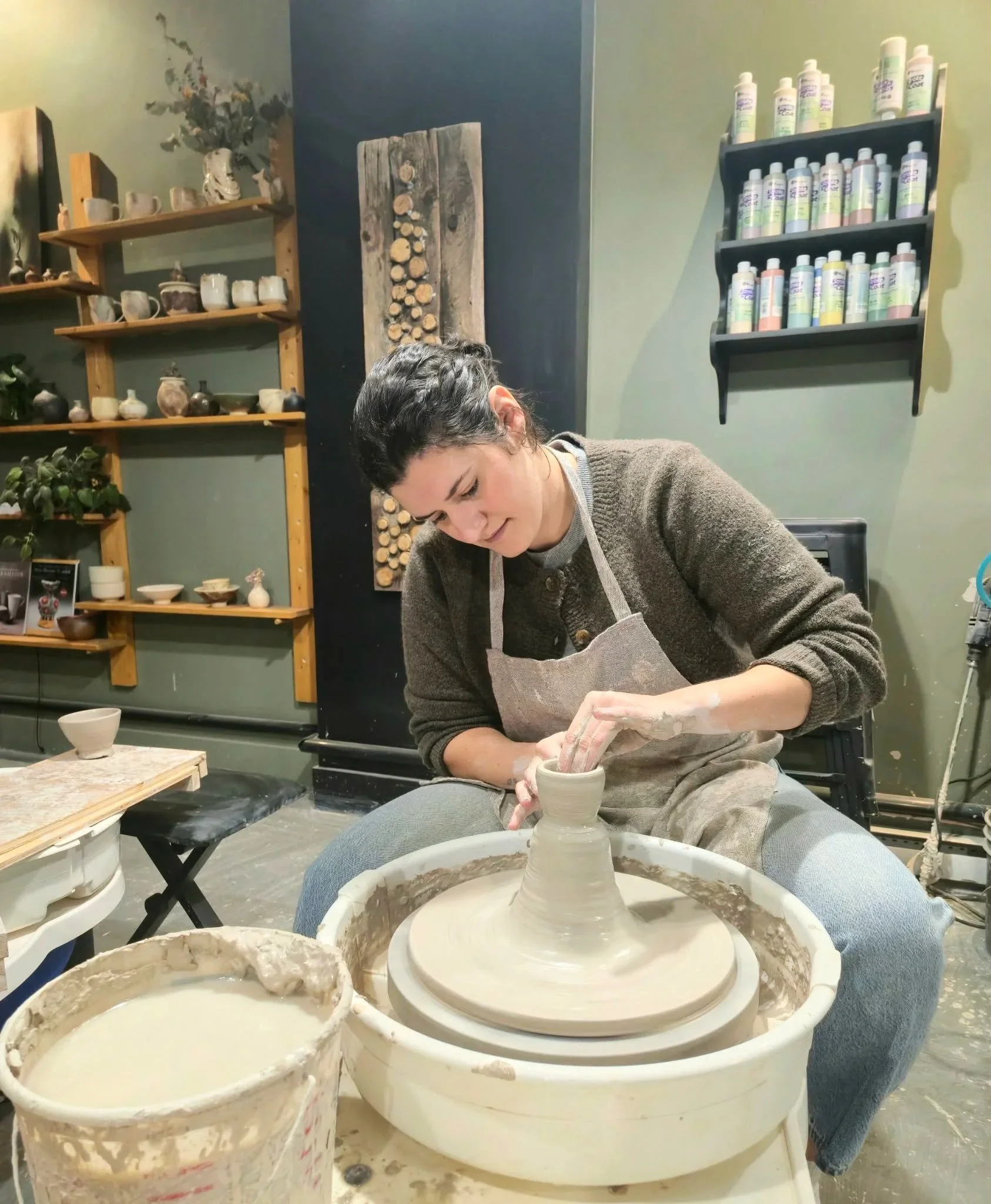 Une femme en train de fabriquer de la poterie sur un tour de poterie dans un atelier. Le fond montre des étagères avec des objets en céramique et des pots de produits.