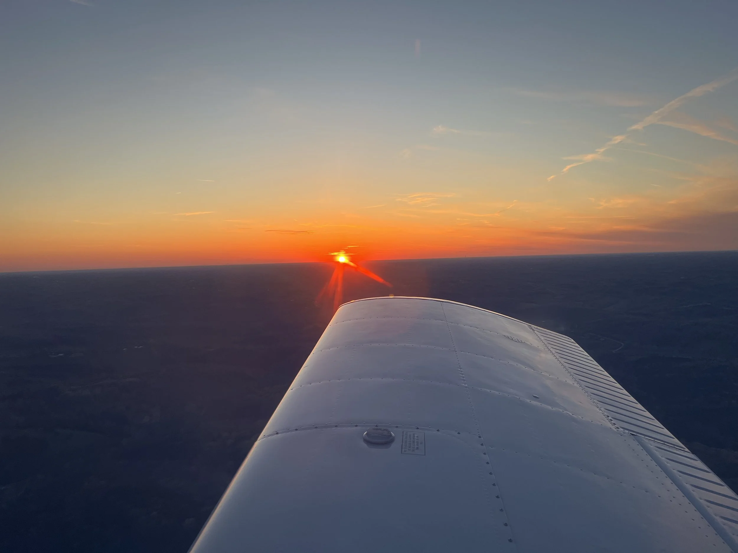 A view from an airplane window during sunset, showing part of the wing and the horizon with the sun setting over the Kentucky landscape.