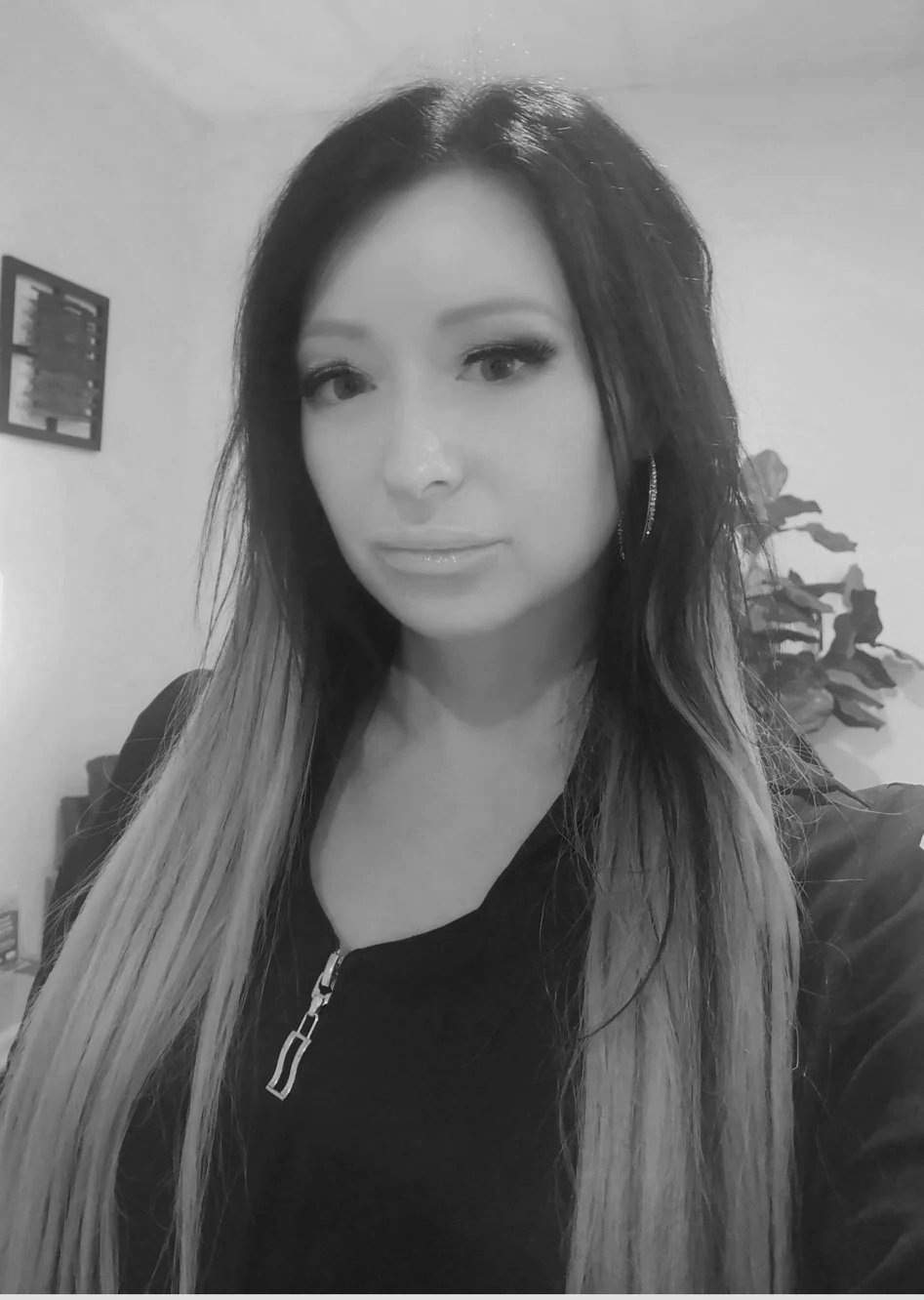 A black-and-white photo of a woman with long, straight hair, wearing a zip-up top with earrings, standing indoors with a plant and a framed picture on the wall behind her.