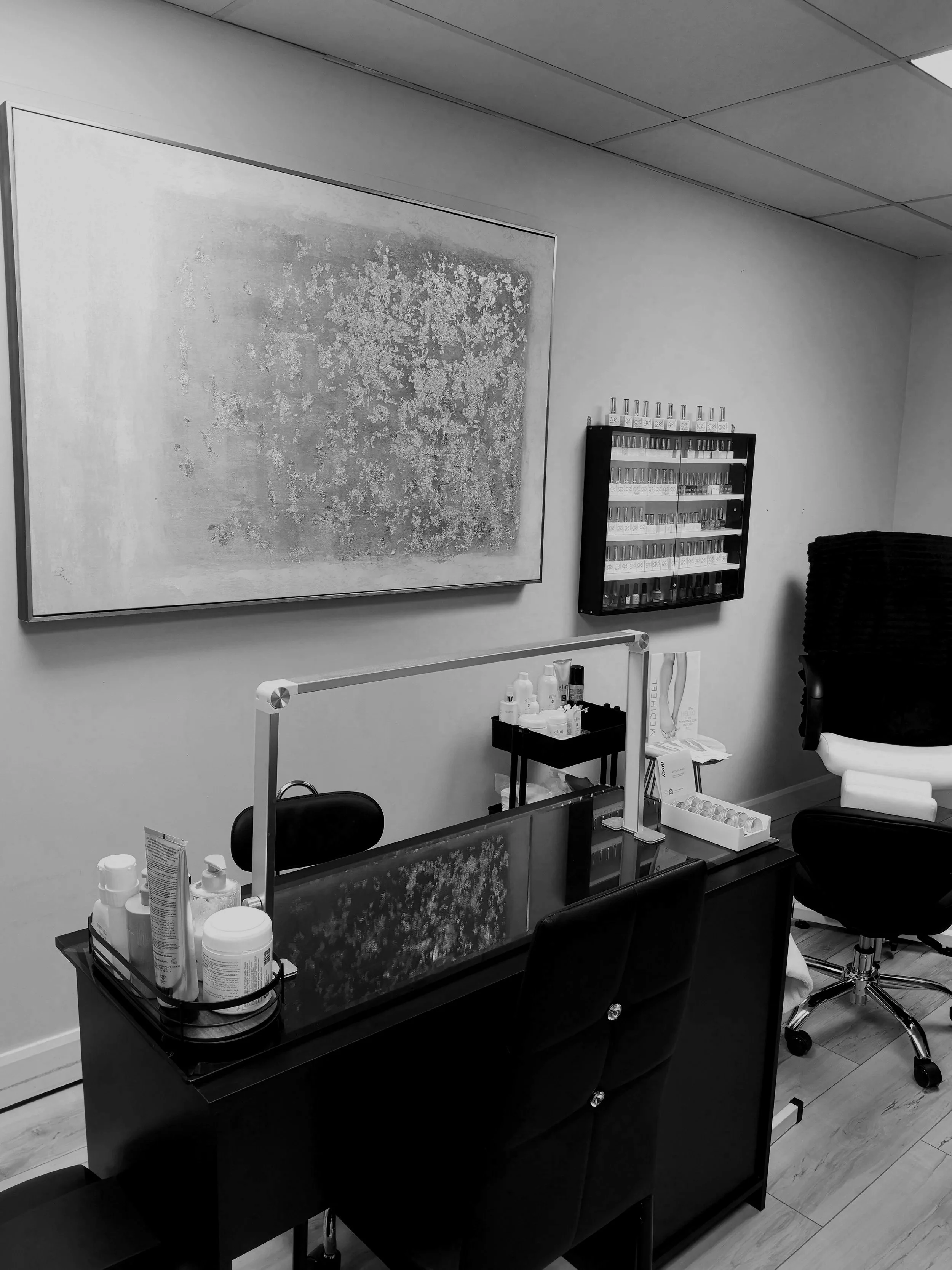 A black-and-white photo of a nail salon treatment room with a workstation, chairs, shelves with nail polish bottles, and an abstract artwork on the wall.