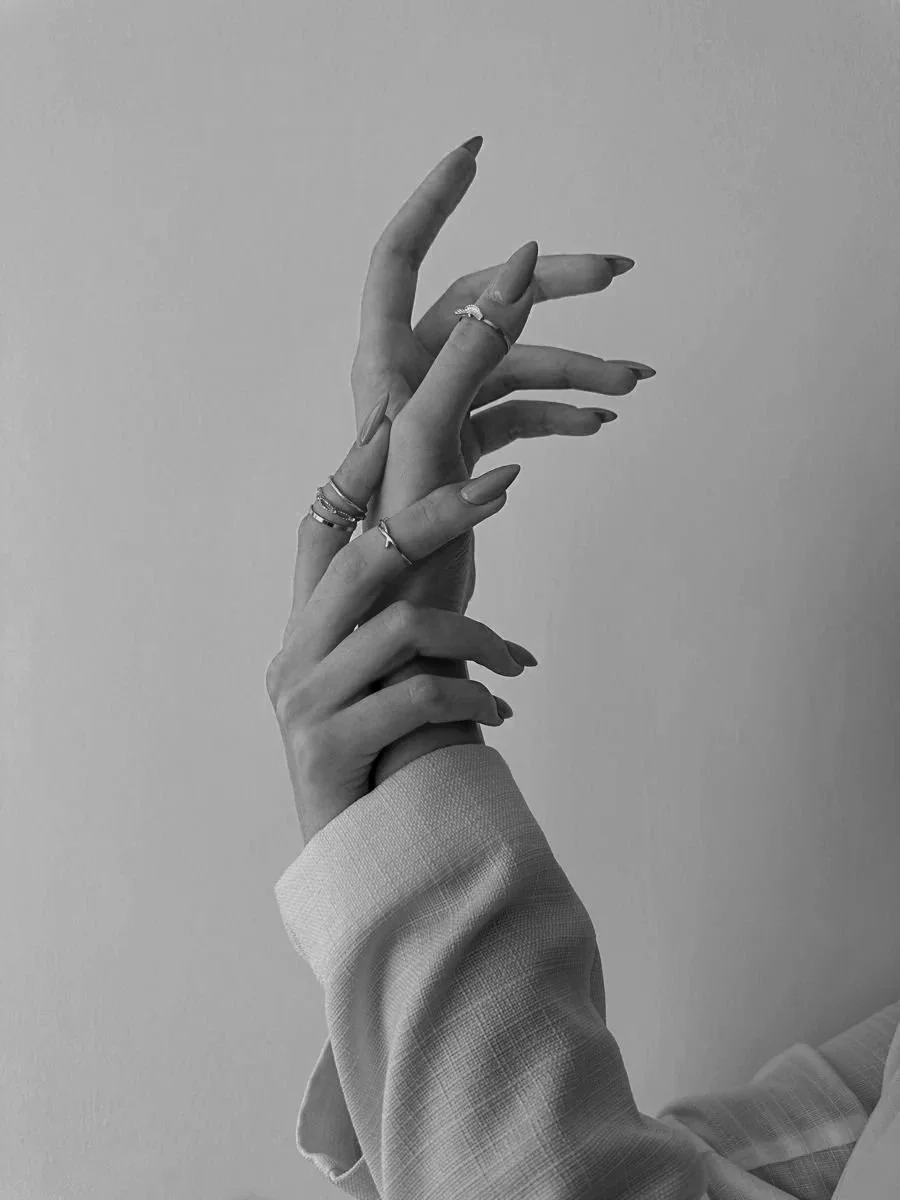 Black and white photo of two hands crossing each other, with rings on some fingers, against a plain background, in a close-up shot.