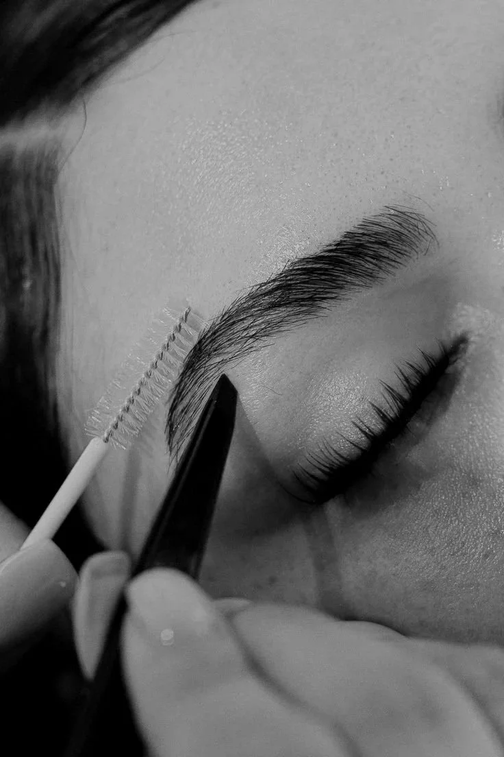 Close-up of a person applying eyelash extensions or makeup to the eyebrow with a small brush, in black and white.