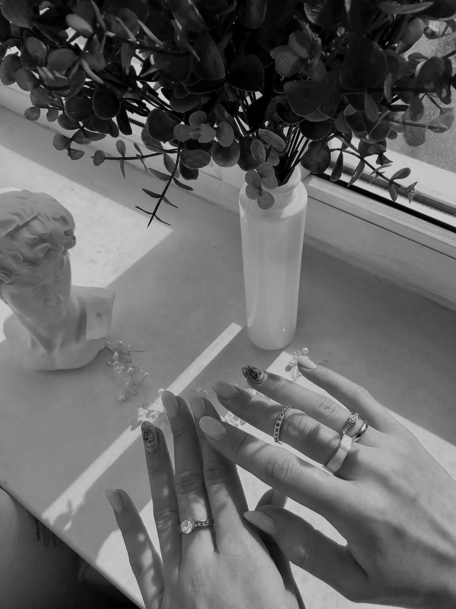 Black and white photo of a person showing off rings on their fingers, with a small bust sculpture, a vase with flowers, and flowering jewelry on a table near a window.