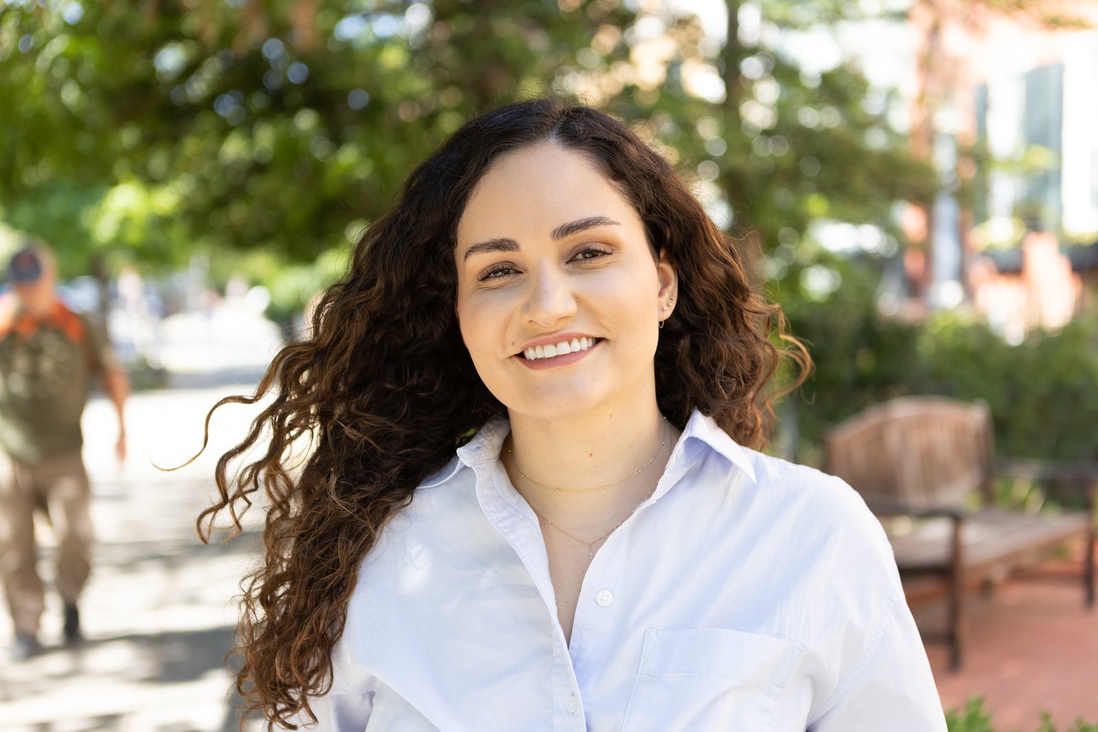 headshot of a smiling woman with curly dark hair wearing a white button-down shirt, photographed outdoors with a blurred green background.