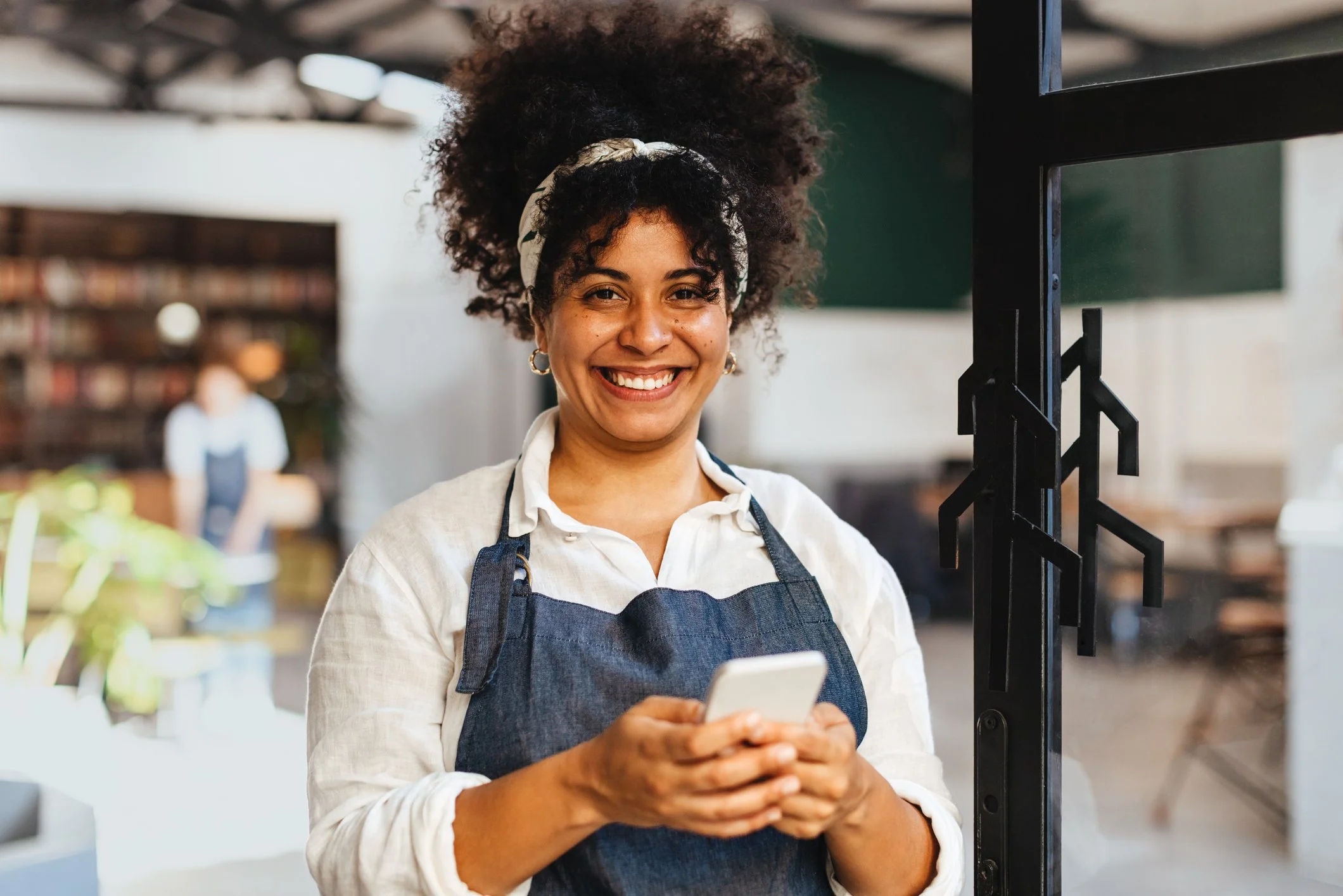 A smiling woman wearing a denim apron holds a smartphone, representing a small business owner or service worker using a mobile app.