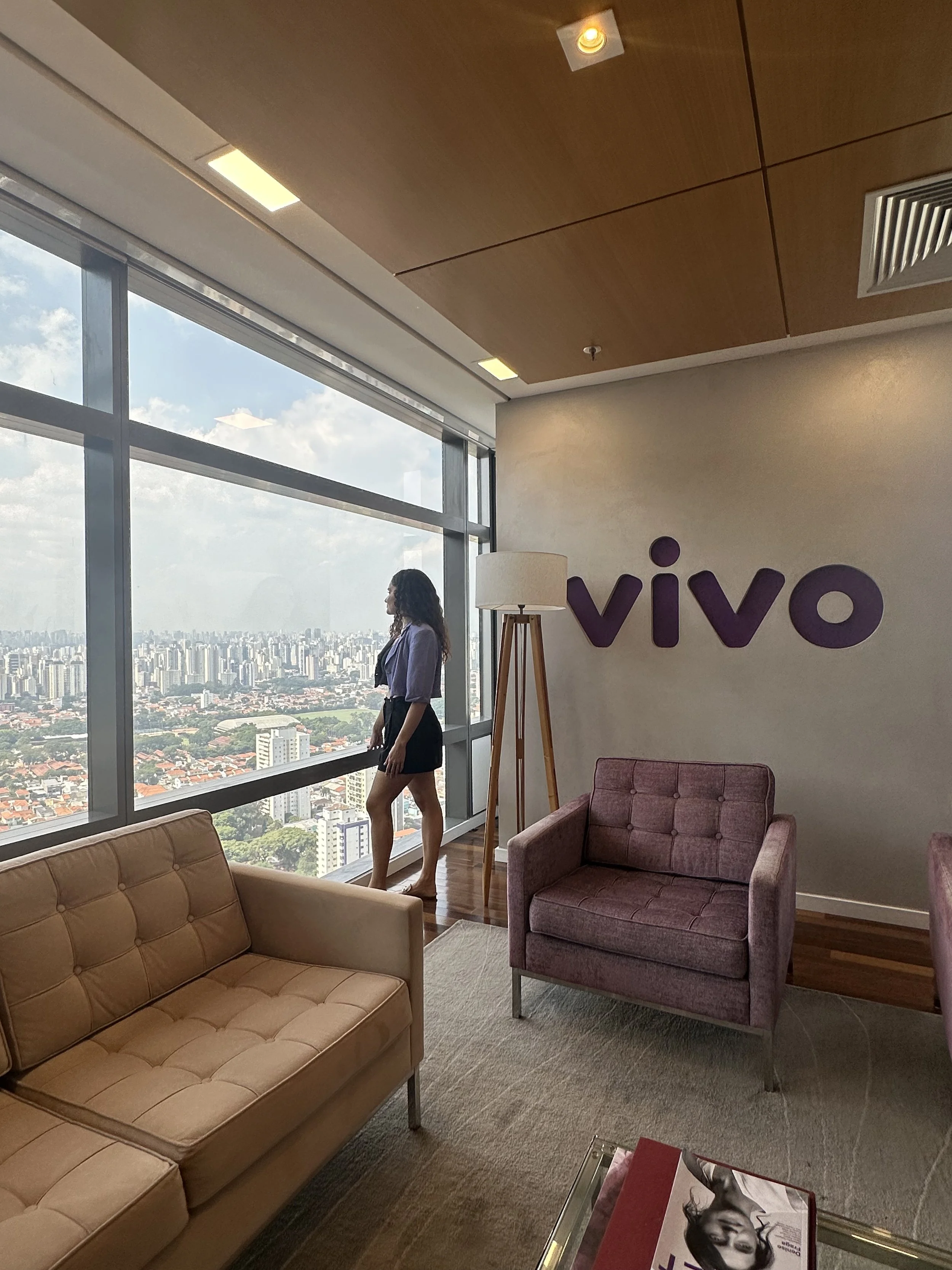 Interior of a modern lounge with large windows showing a city skyline, beige and purple armchairs, a glass coffee table with magazines, a standing lamp, and a wall sign that says 'vivo' with a woman standing near the window.