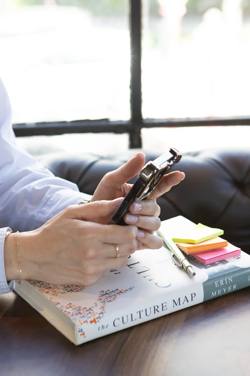 Close-up of a woman's hands holding a smartphone, resting on a copy of The Culture Map by Erin Meyer, with colorful sticky notes and a pen on a table.