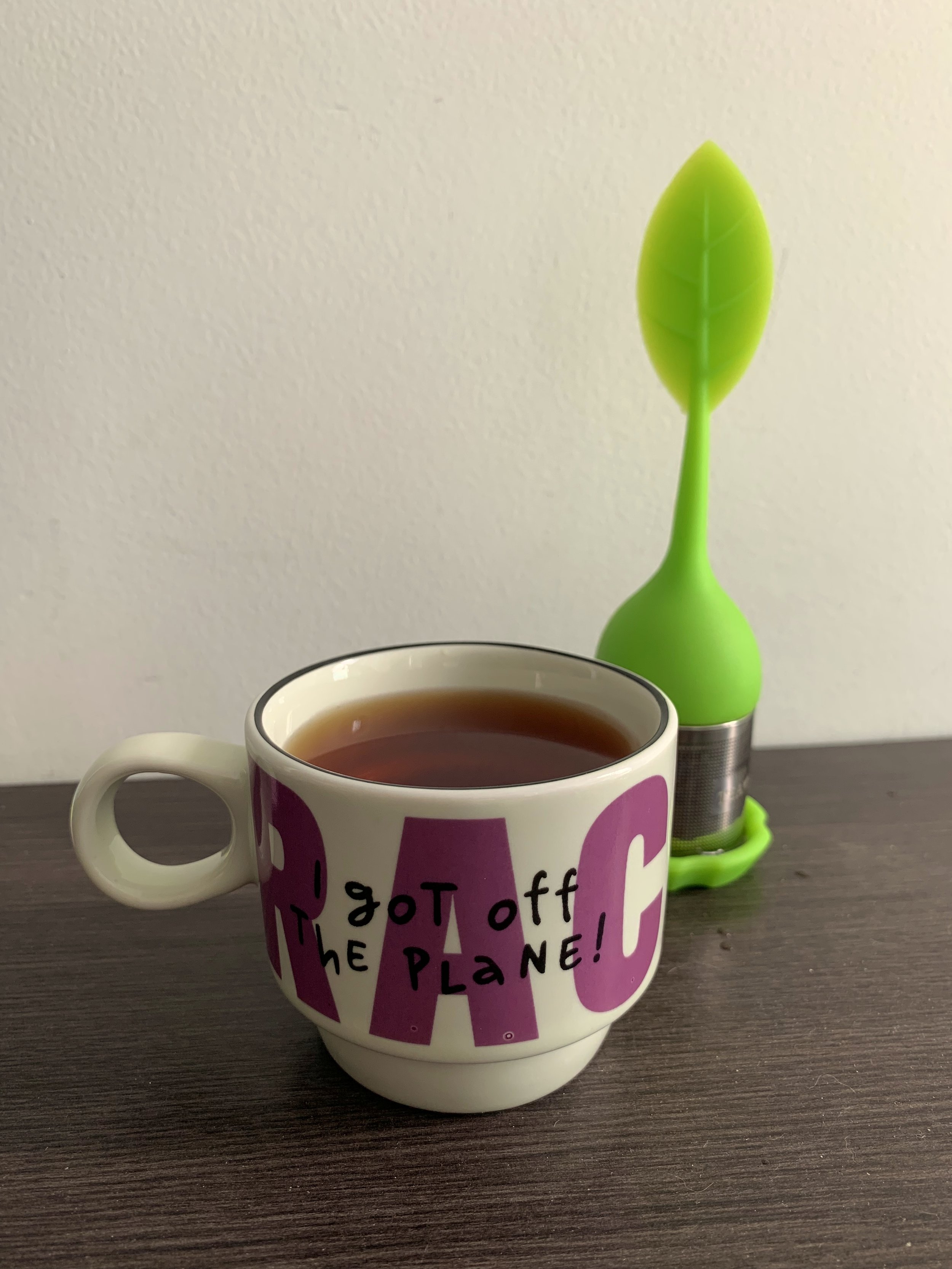 A white ceramic mug with purple lettering filled with tea, next to a green leaf-shaped silicone tea infuser resting on a wooden surface.