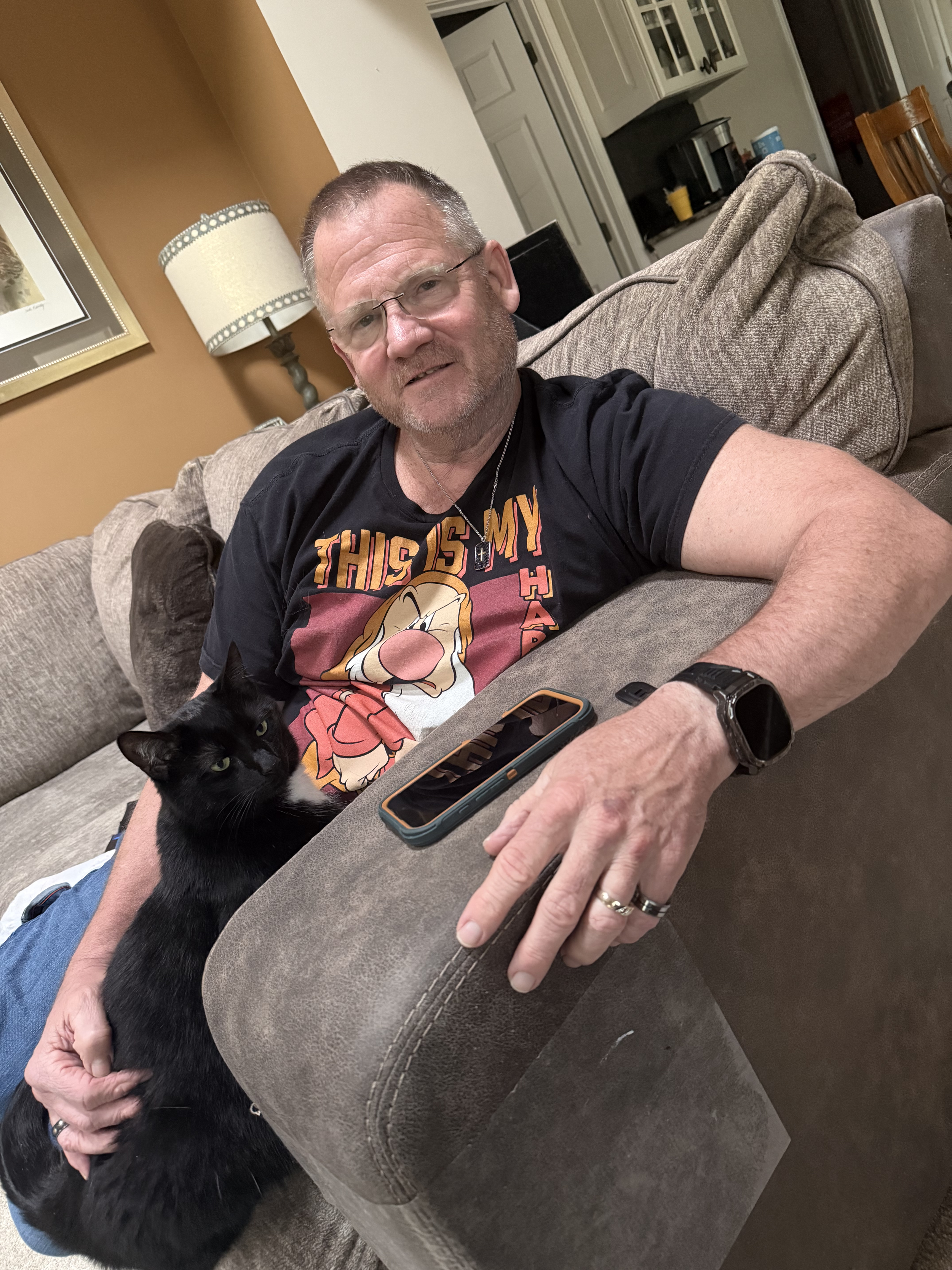 A man sitting on a brown couch, wearing glasses, a black T-shirt with a cartoon character, and a watch, holding a black and white cat on his lap. The room has a lamp, framed artwork, and a kitchen in the background.