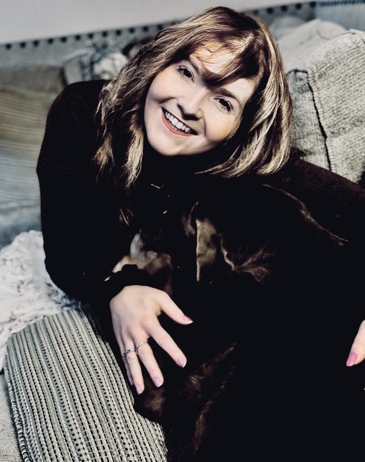 A woman with shoulder-length light brown hair smiling and holding a dark brown dog with black and tan markings on a beige and brown striped couch.