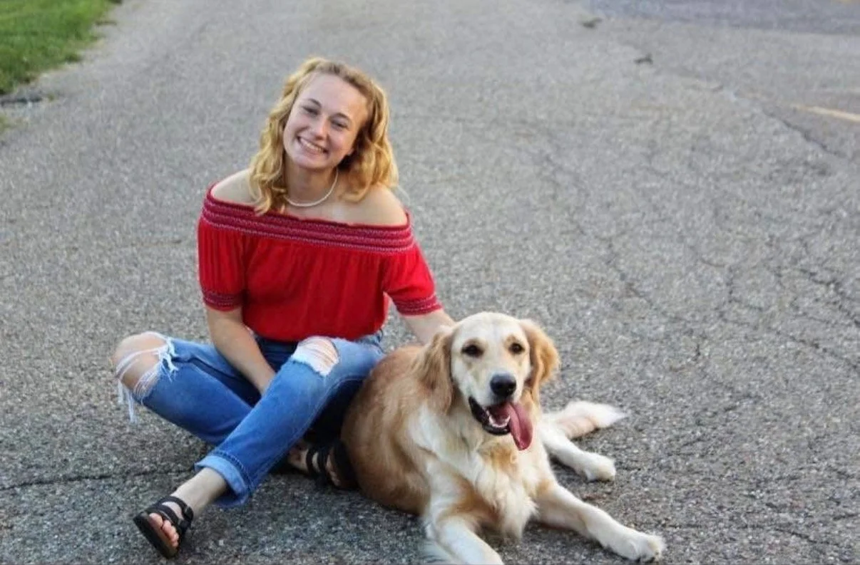 A young woman with blonde curly hair sitting on an asphalt road next to a golden retriever dog, both smiling at the camera.