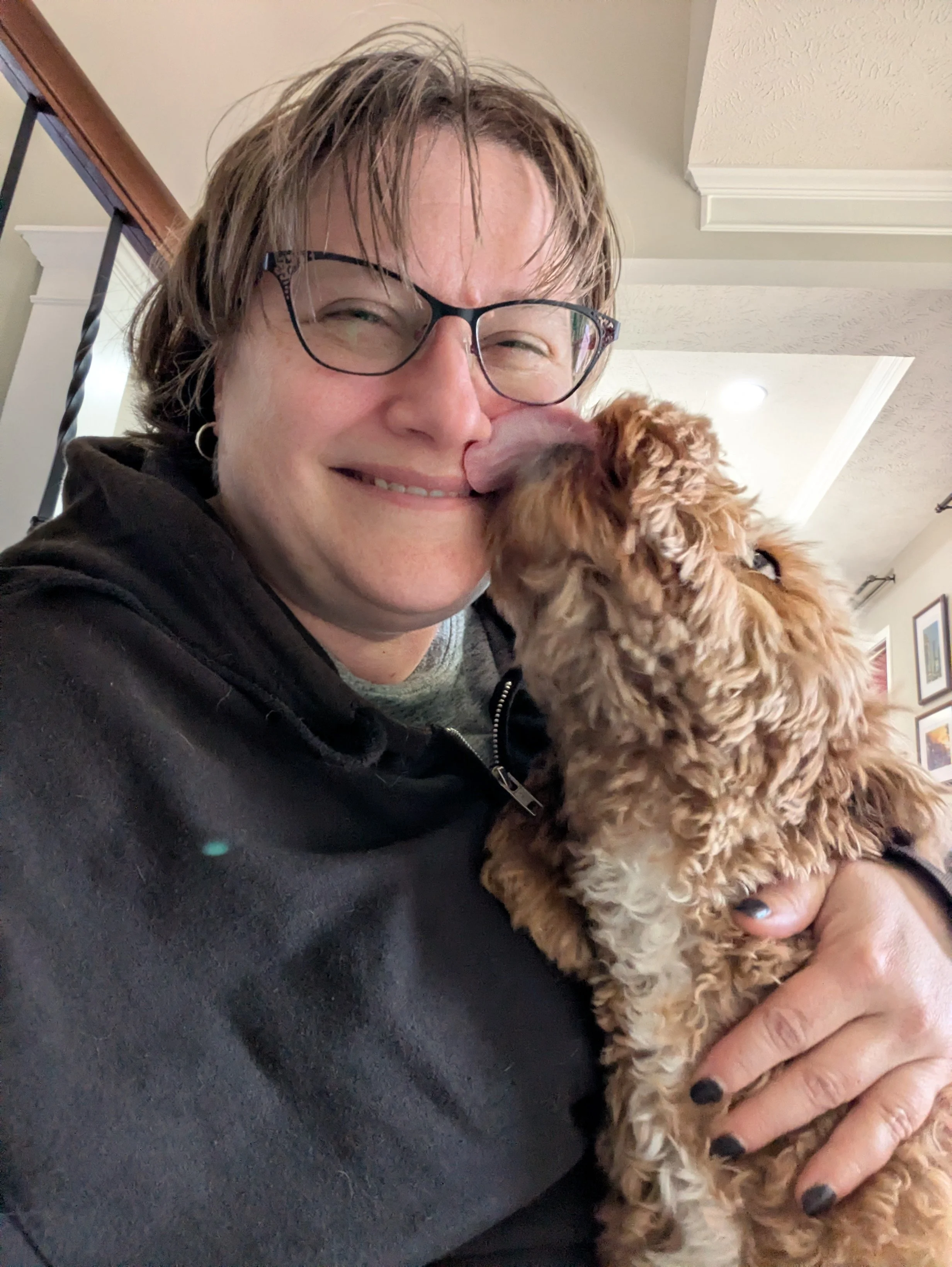 A woman with glasses smiling while holding an affectionate light brown, curly-haired dog.