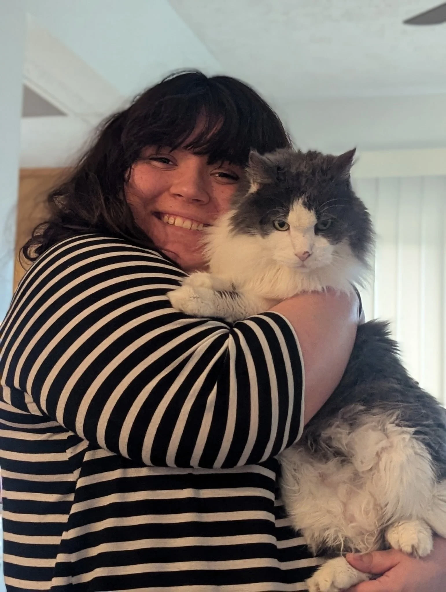 Person with dark hair and striped shirt holding a large, fluffy black-and-white cat indoors near a window and ceiling vent.