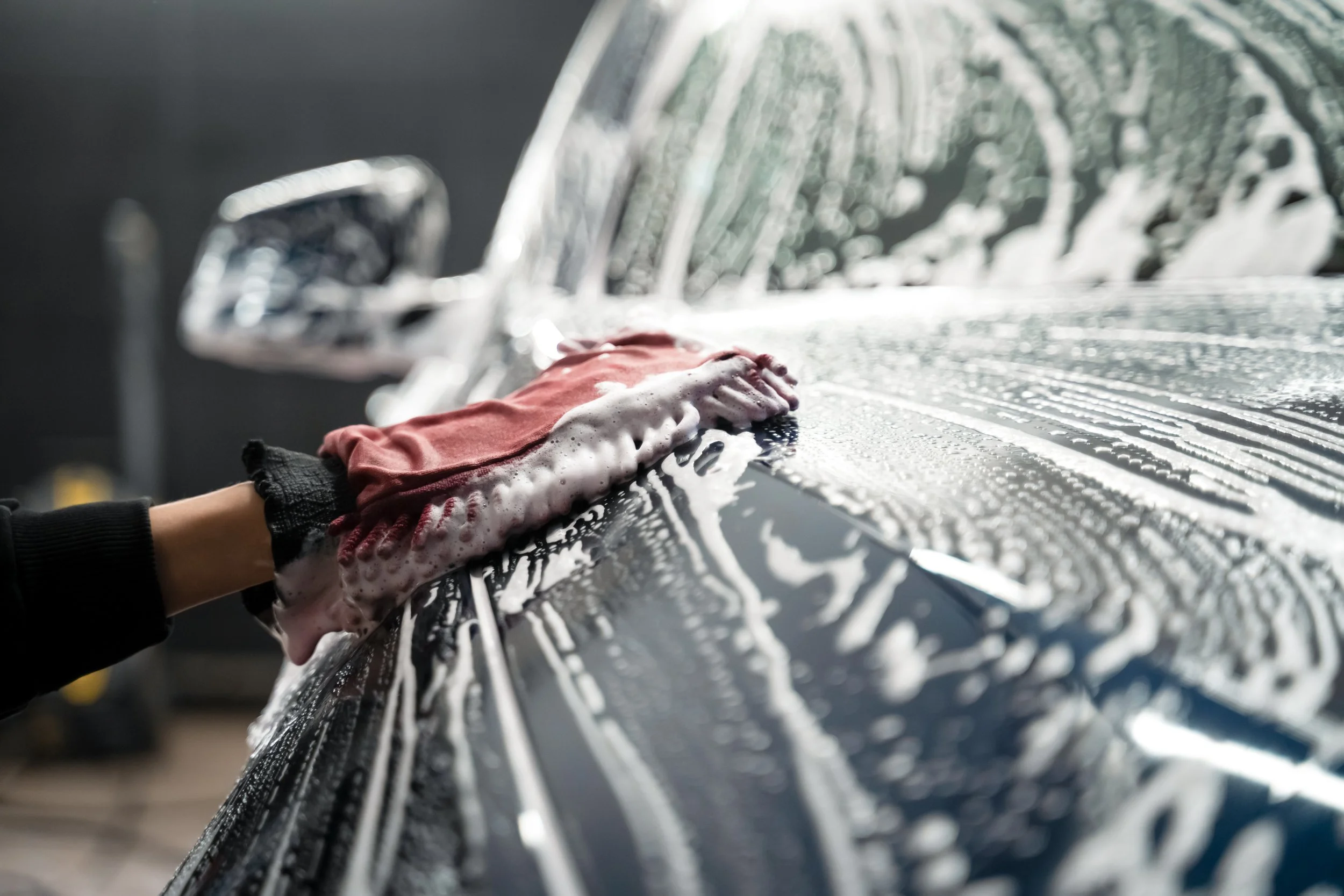 A person wearing a black long sleeve shirt and a black glove is washing a dark-colored car with soapy water and a pink cloth.