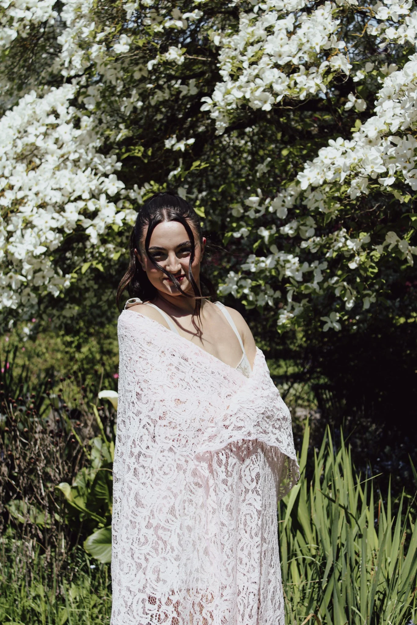 A woman with dark hair styled in an updo with braids, wearing a white lace dress, standing outdoors surrounded by green plants and white flowering bushes.
