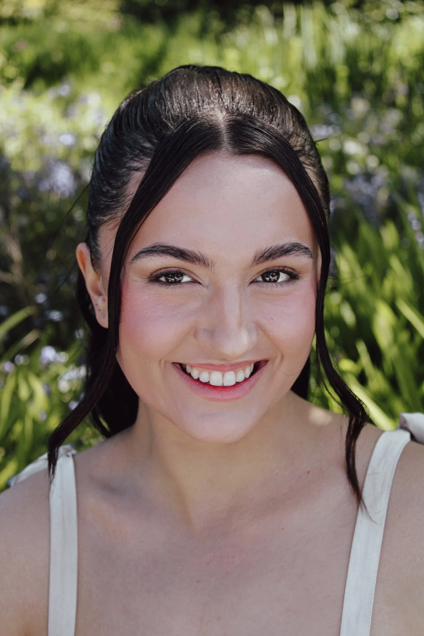 Close-up of a smiling woman with dark hair and white tank top, outdoors with green foliage in the background.