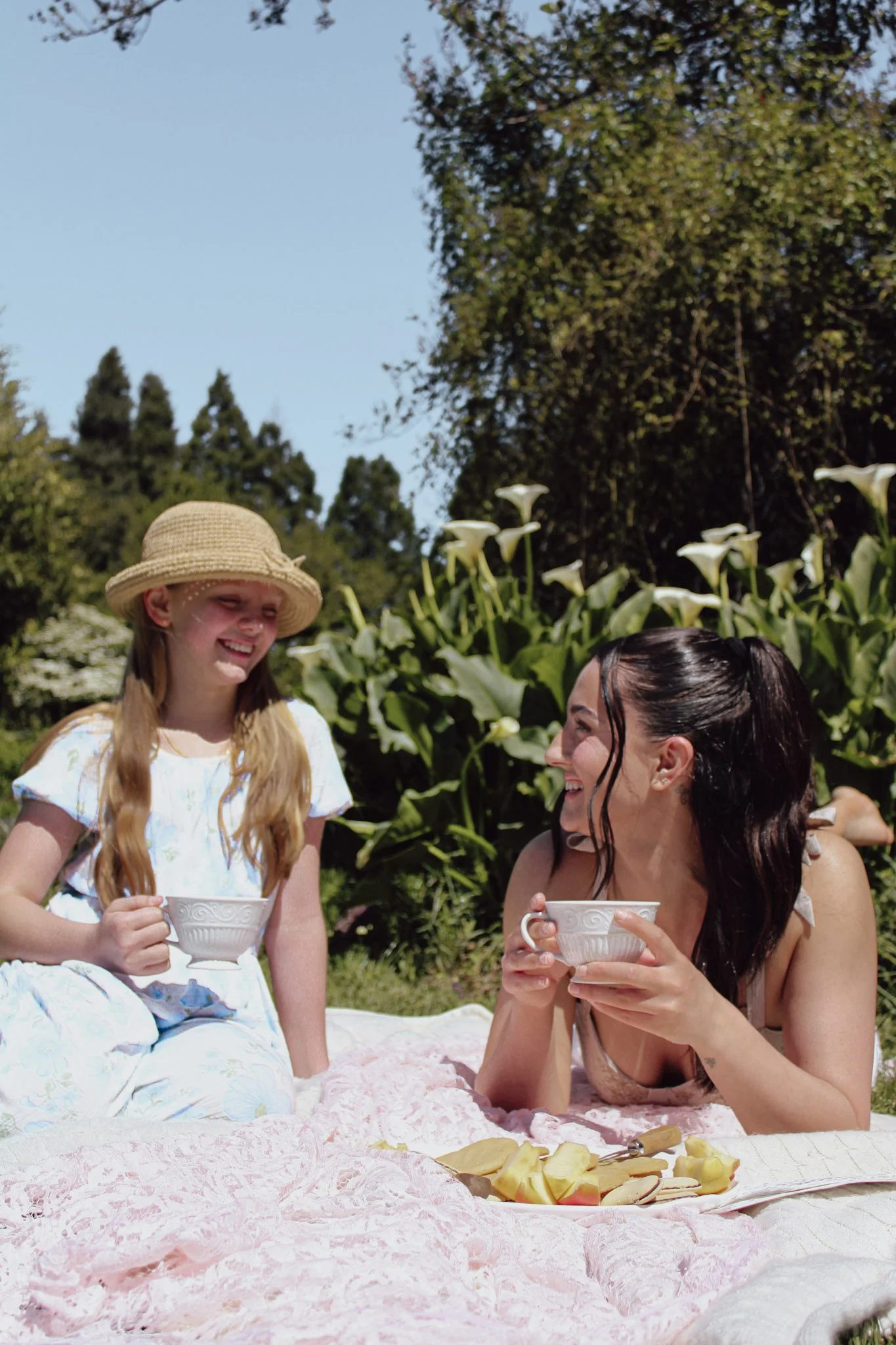 Two women and a girl sitting on a pink blanket outdoors, having a tea party with cups and fruit, surrounded by greenery and flowers under a blue sky.