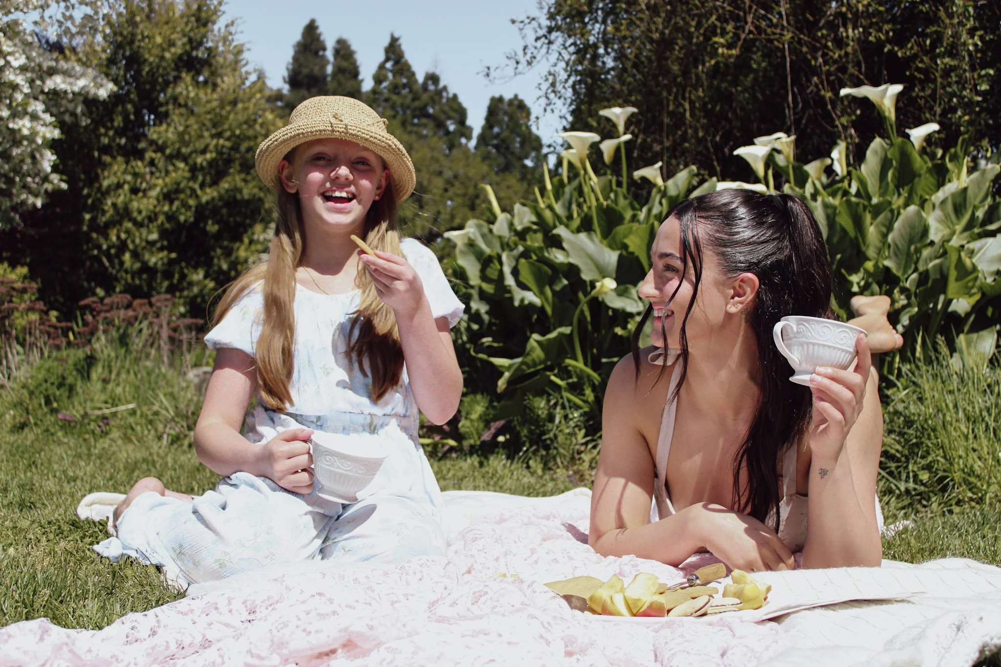 A young girl with blonde hair wearing a straw hat and a white dress, sitting on a blanket and smiling, while an older woman with wet dark hair and a white dress, smiling and holding a white bowl, sit outdoors among greenery and blooming flowers durin
